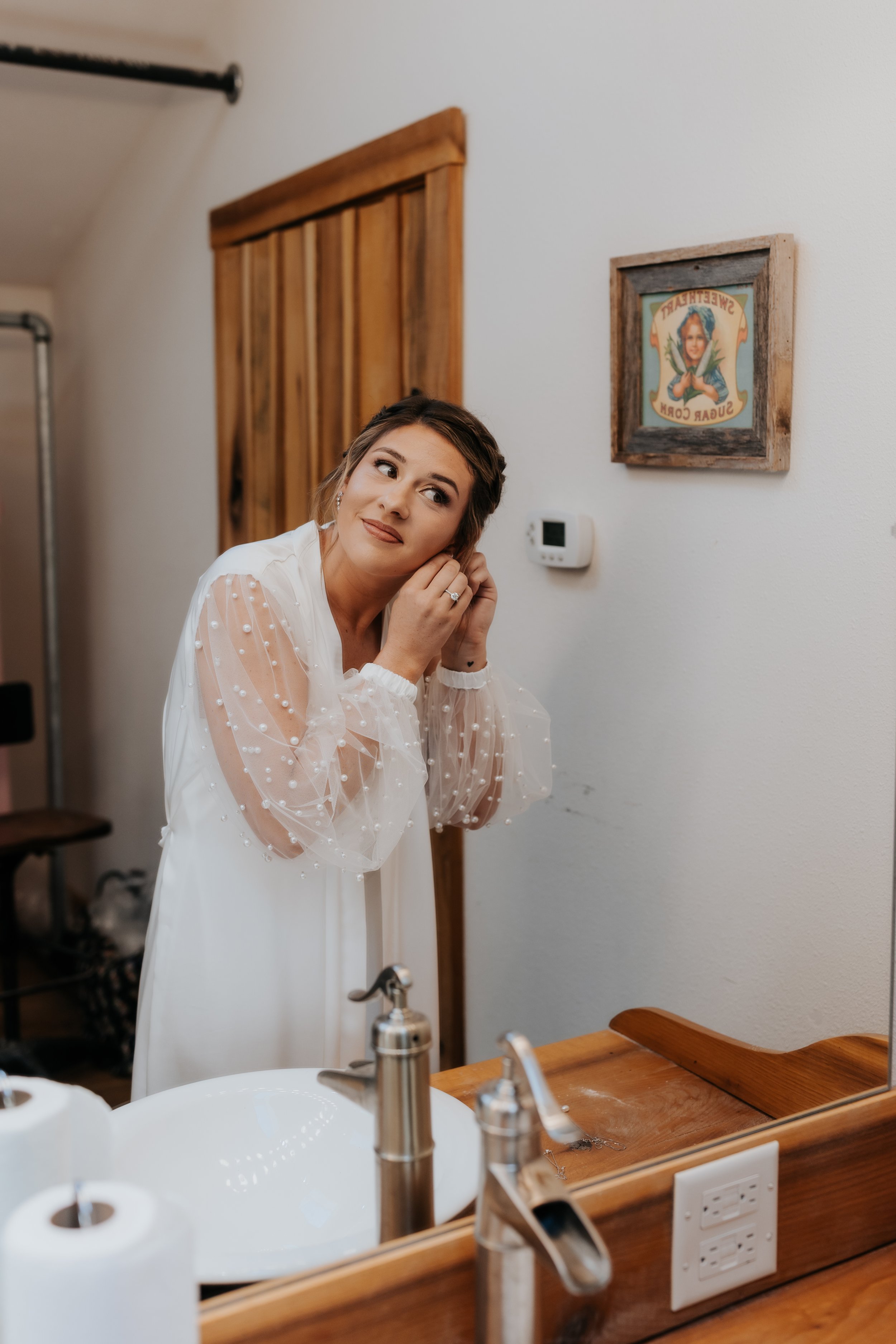 A woman in a white dress with sheer sleeves is looking into a bathroom mirror, adjusting her earring, in a room with light-colored walls, wooden accents, and a framed picture on the wall.