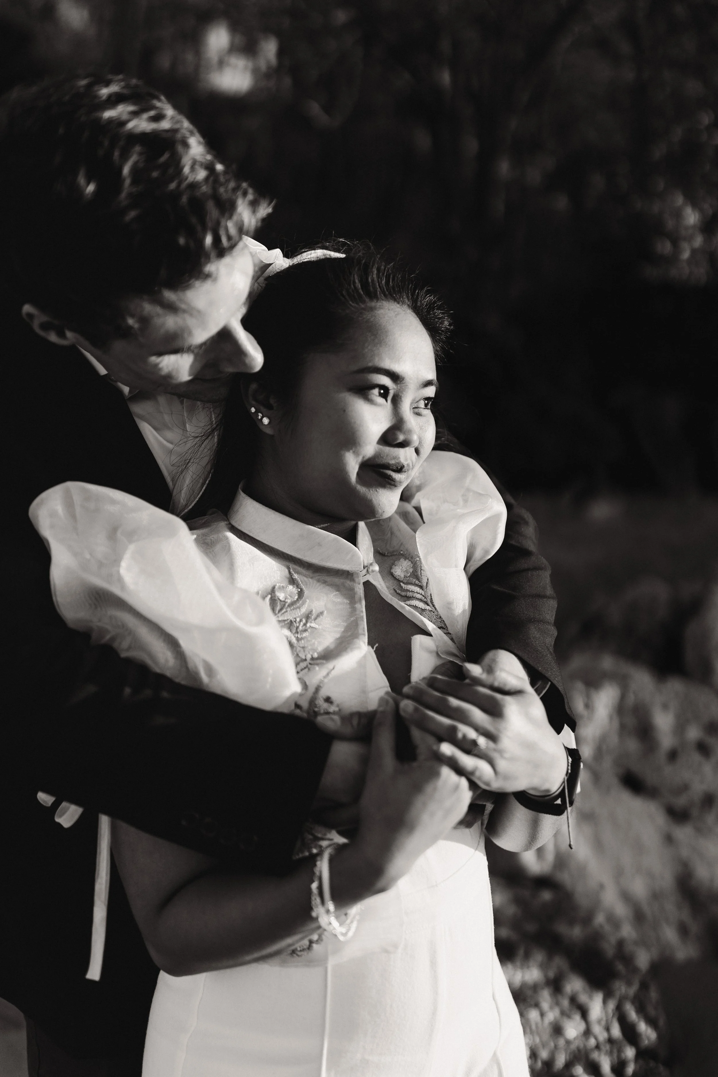 A black and white photo of a couple embracing outdoors at night. The woman has her hands crossed over her chest, and the man is leaning close to her, with only part of his face visible.