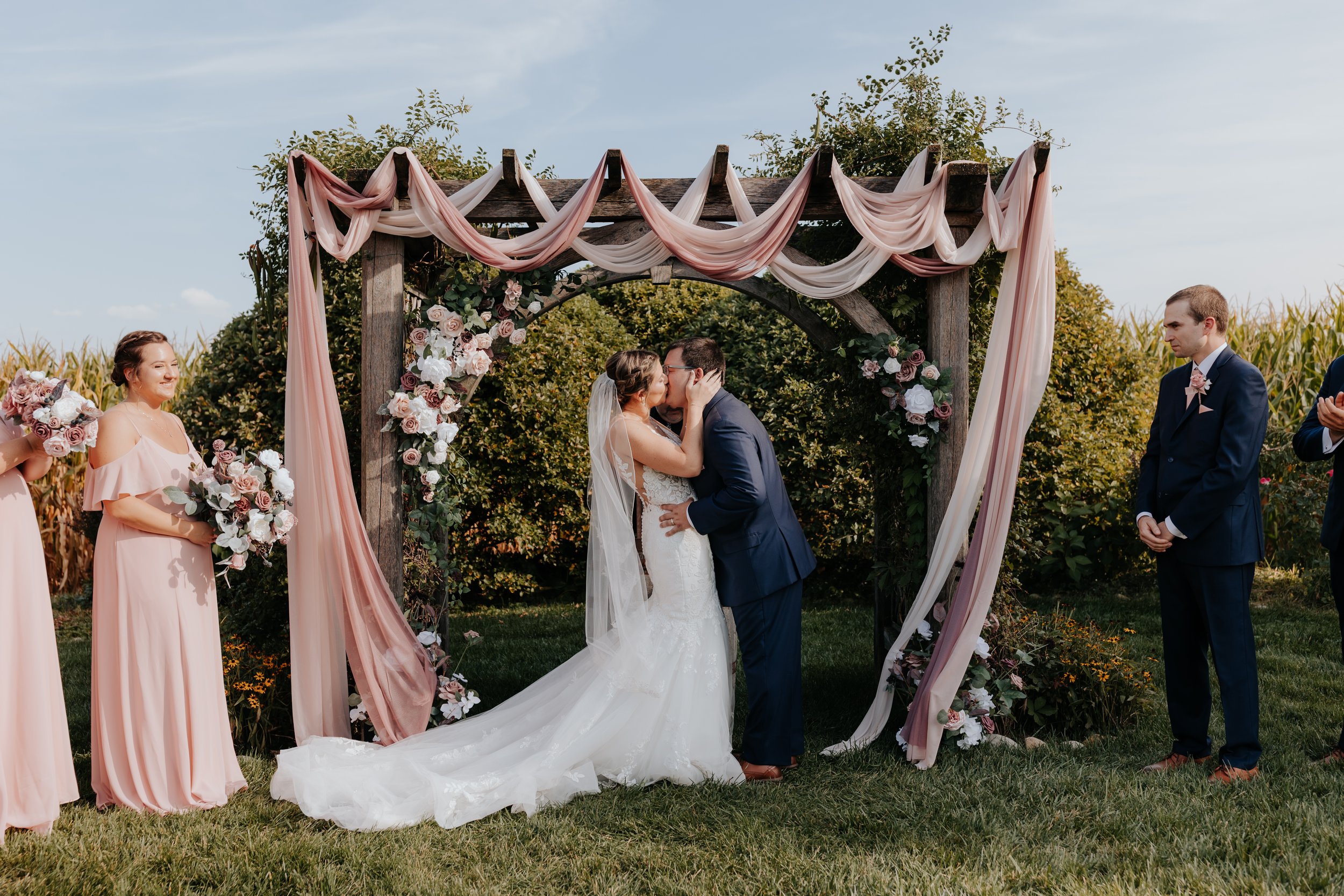 A bride and groom kiss under a wedding arch decorated with pink and white drapes and flowers, with bridesmaids in pink dresses holding bouquets on the left and groomsmen in navy suits on the right, outdoors on a grassy field during daytime.