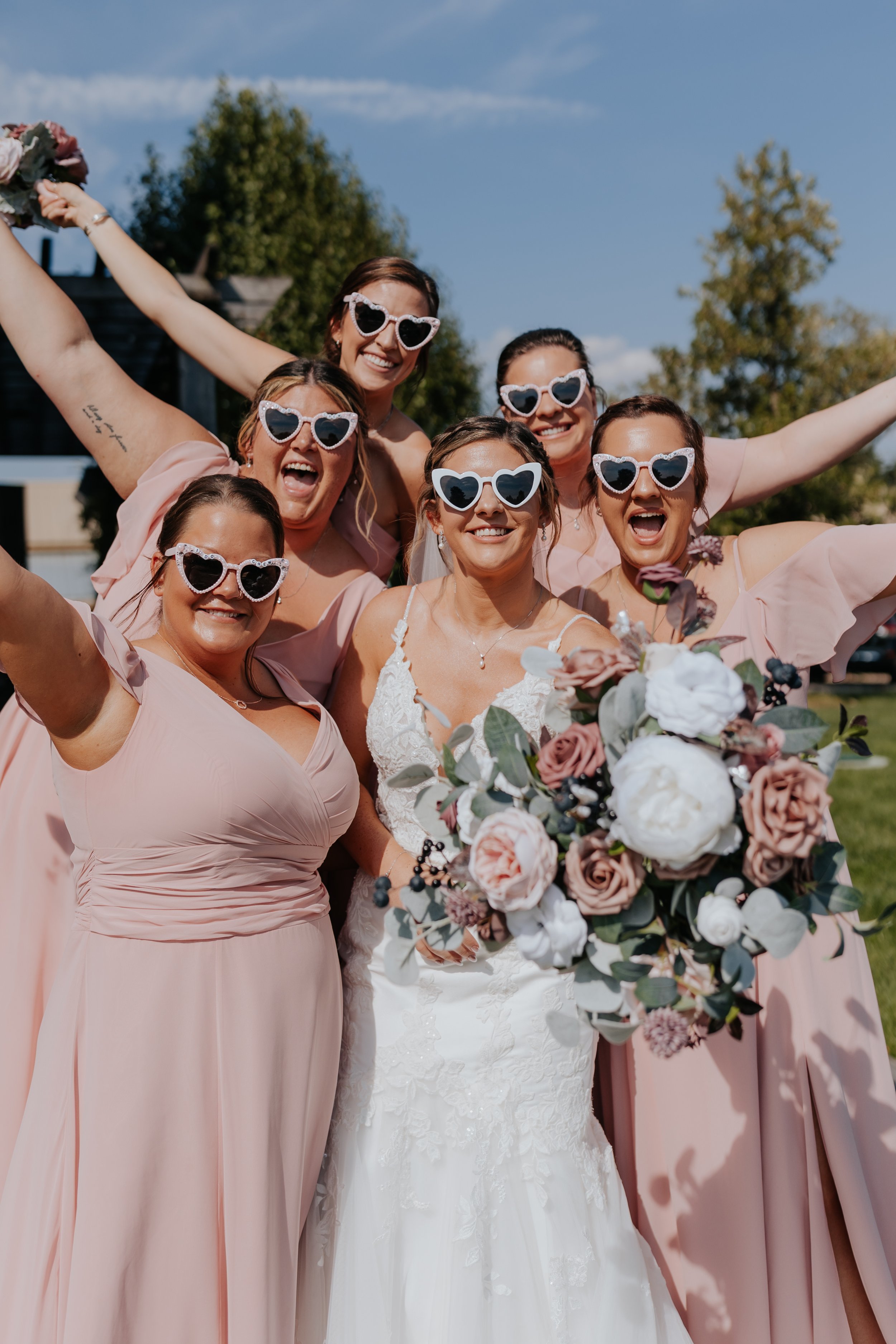 A bride with a bouquet of flowers and six bridesmaids in pink dresses, all wearing matching heart-shaped sunglasses, celebrating outdoors on a sunny day.