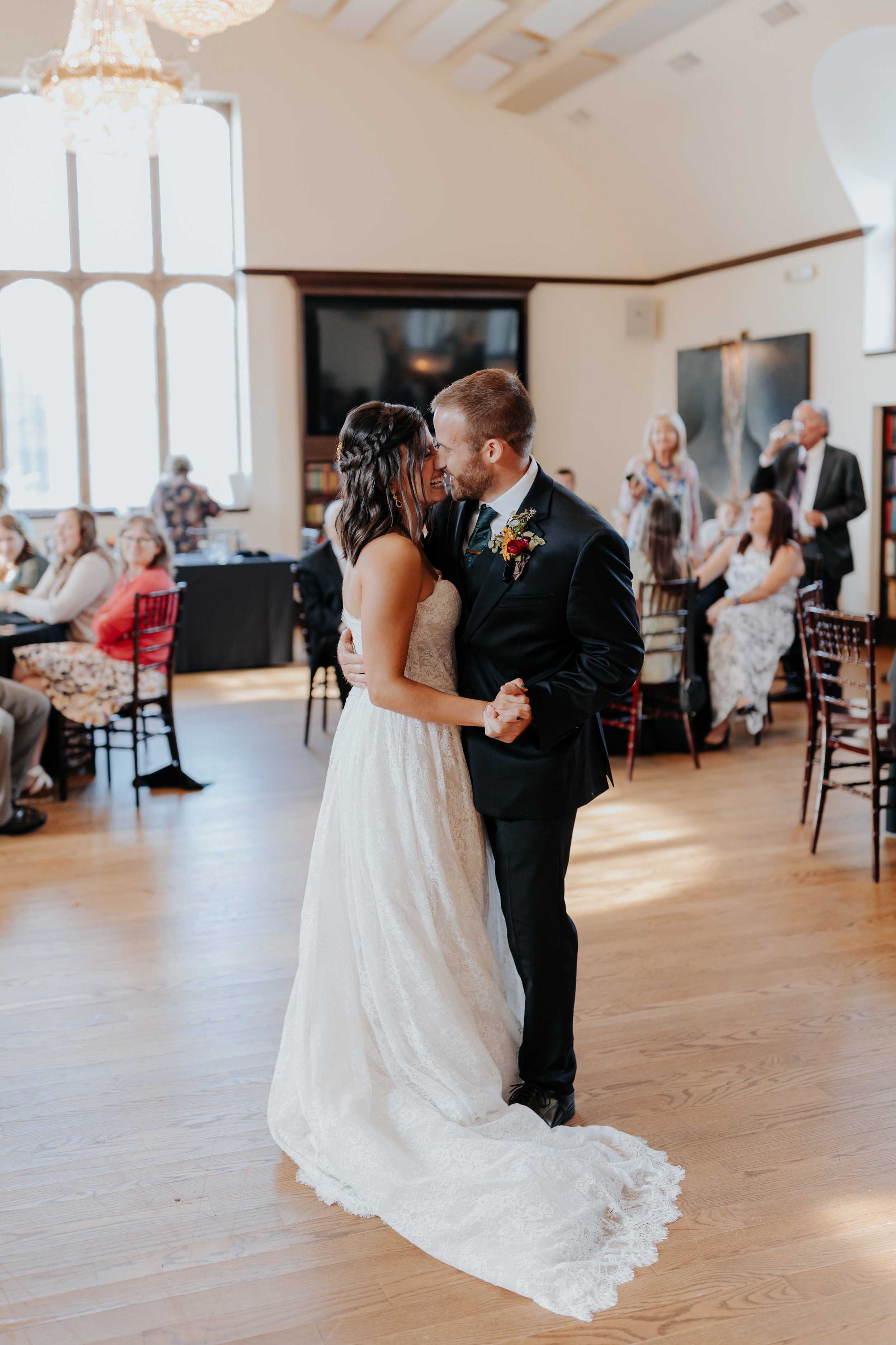 A newlywed couple shares a dance at their wedding reception, surrounded by seated guests in a bright, elegant venue.