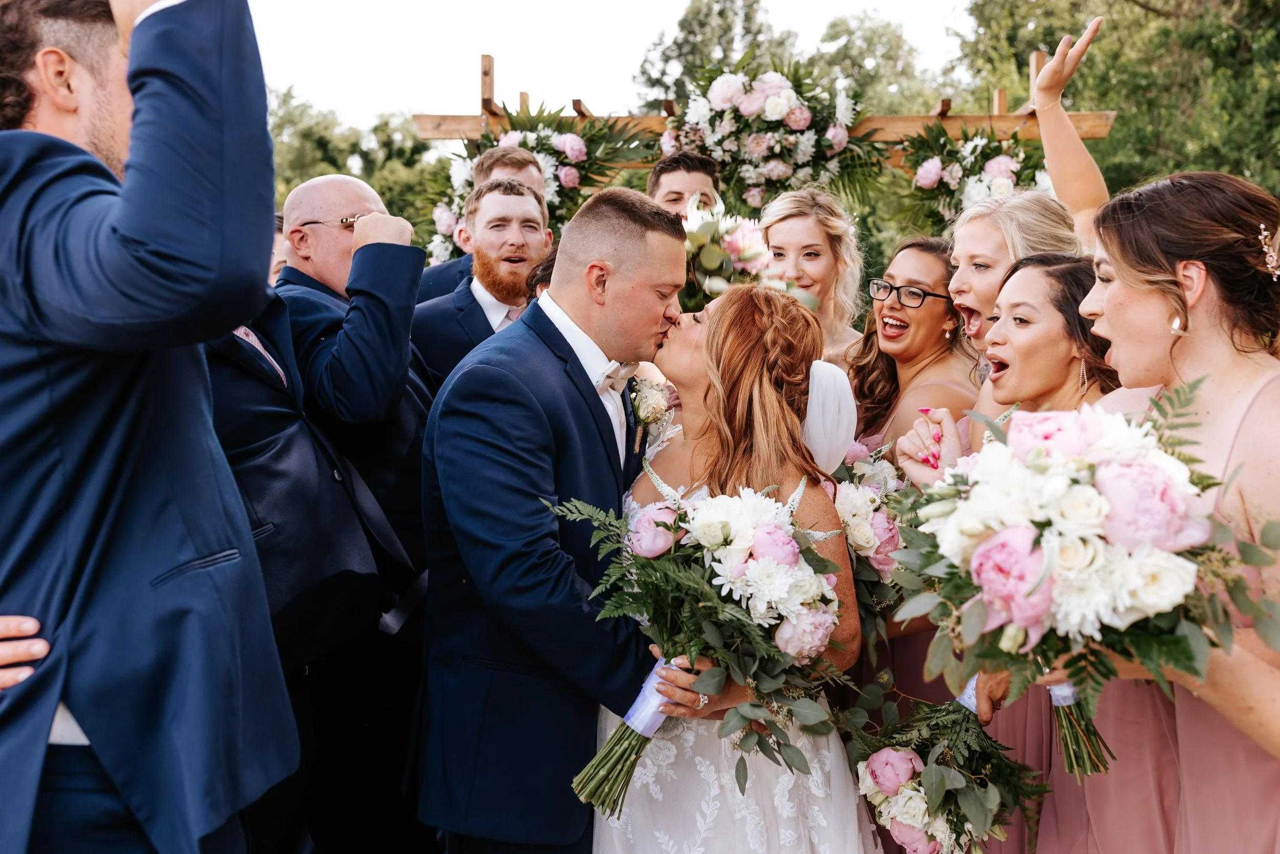 A bride and groom kissing during their wedding ceremony surrounded by friends and family holding bouquets of pink and white flowers, outdoors under a floral arch.