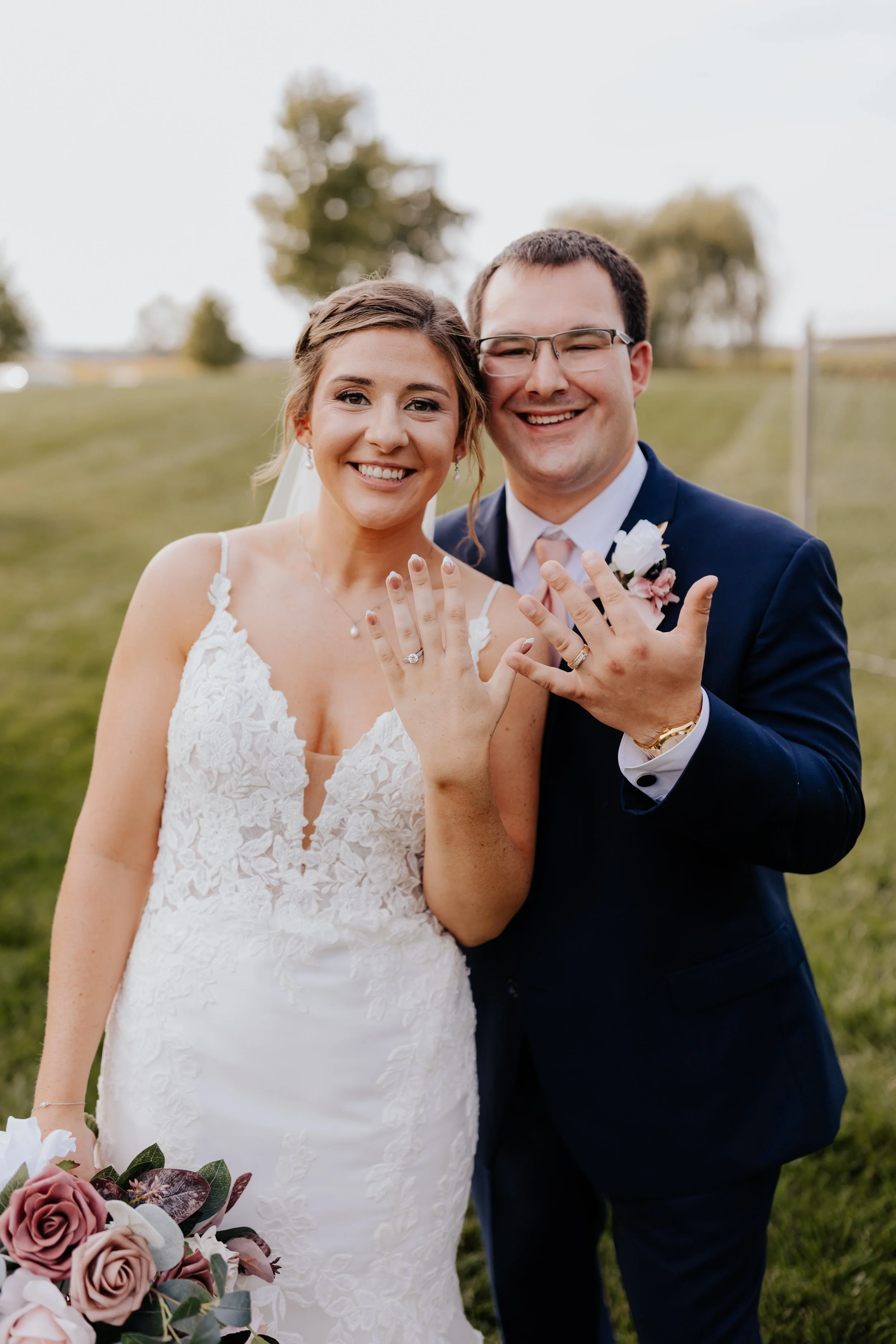 A newlywed couple outdoors showing off their wedding rings, with the bride in a white lace wedding dress and the groom in a navy blue suit, smiling at the camera, holding hands, and standing on a grassy field.