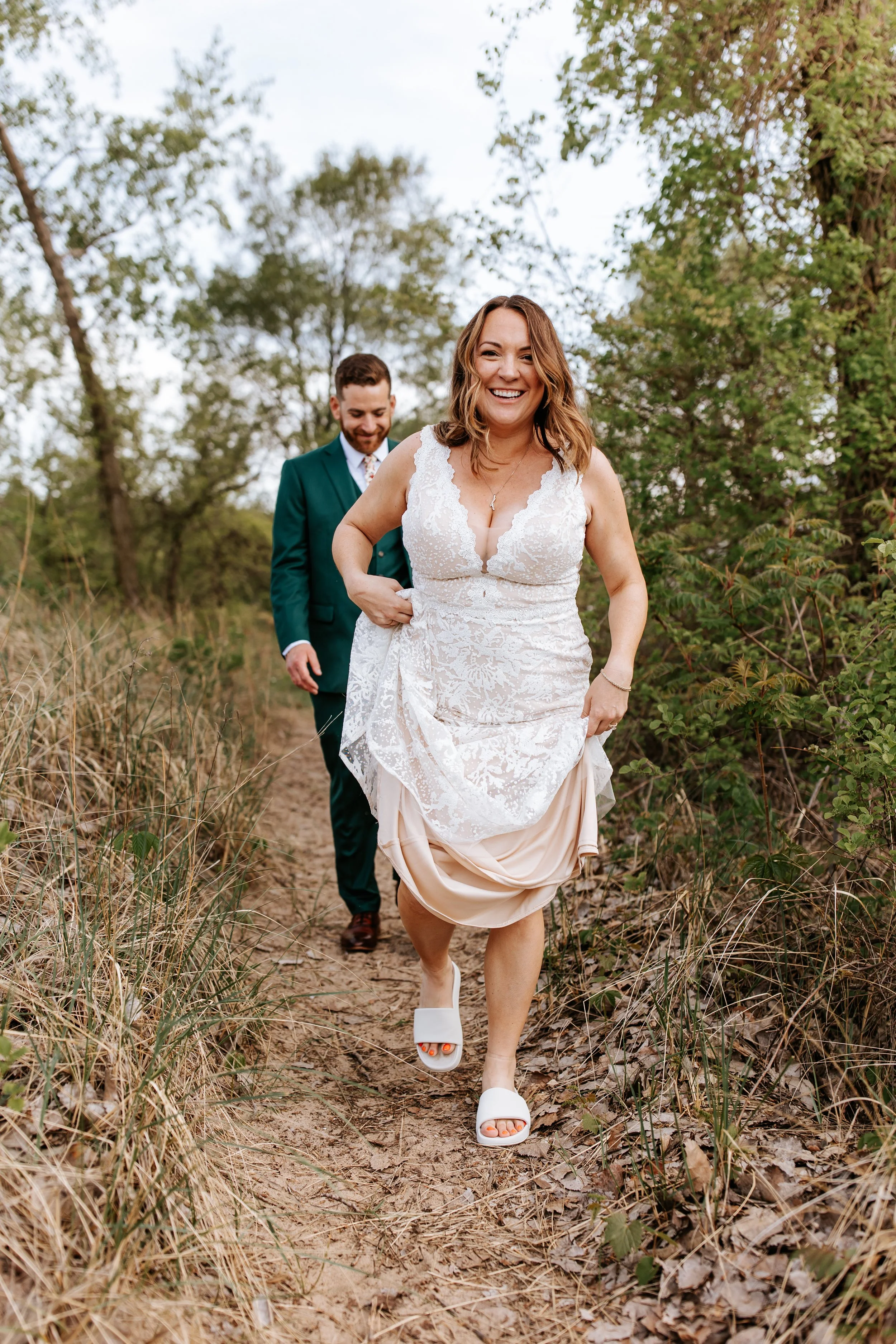 A woman in a white lace wedding dress and a man in a green suit walking on a dirt path through a wooded area, smiling happily.