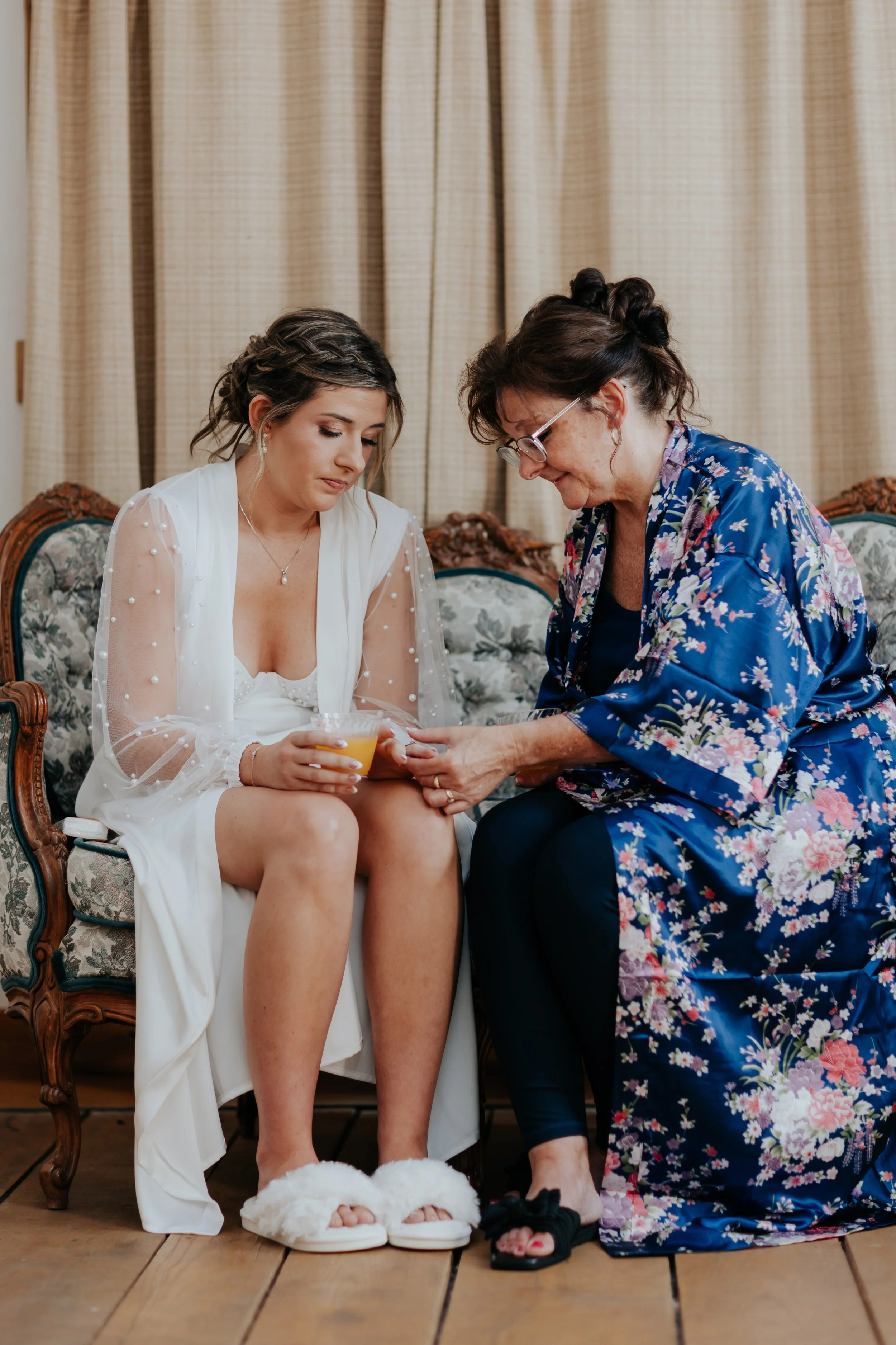 A bride sitting on a vintage sofa, wearing a white robe and slippers, holding a glass of orange juice. An older woman, possibly her mother, is kneeling beside her, holding her foot and wrapping it with a bandage. The setting appears to be in a cozy r