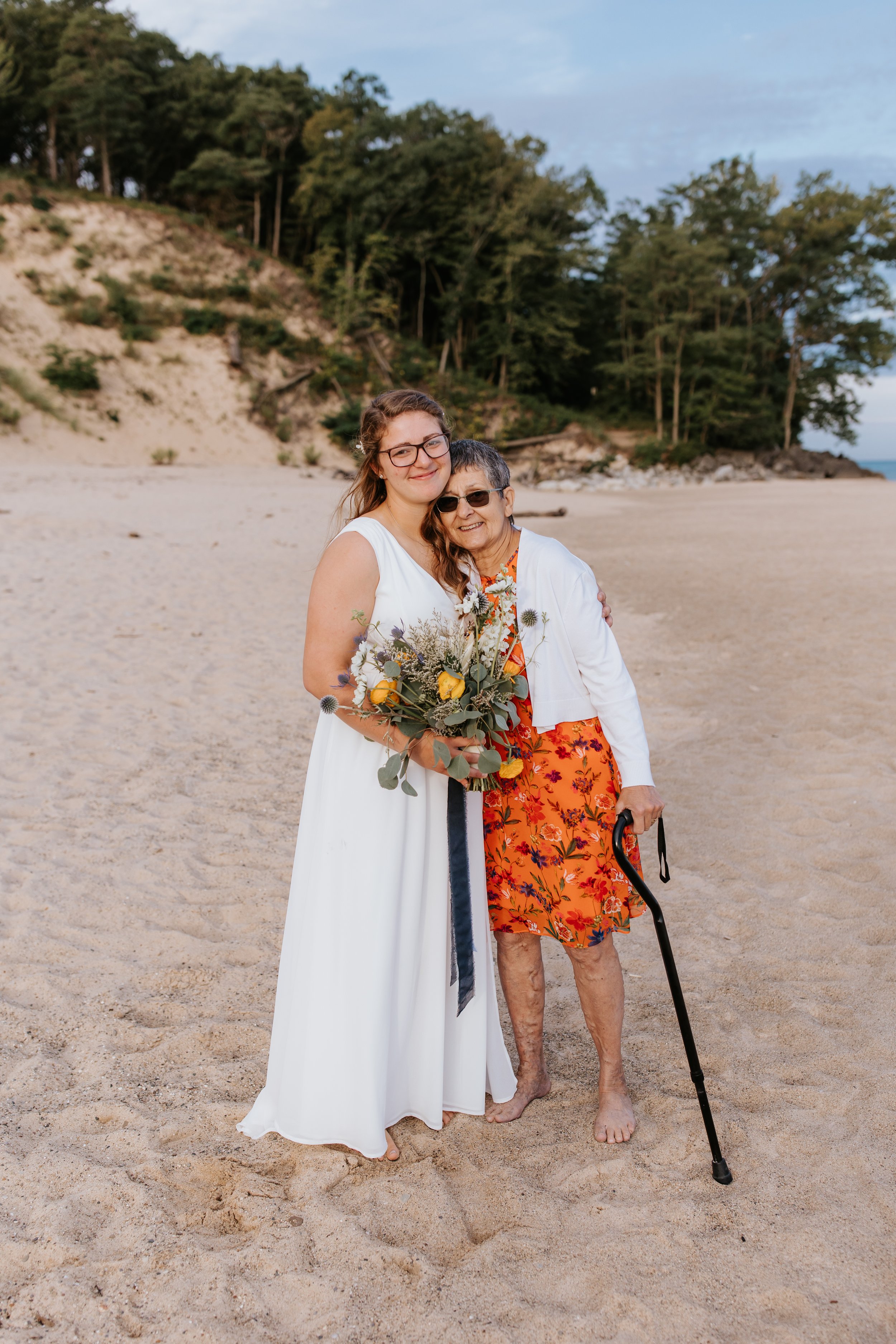 A young woman in a white dress holding a bouquet of flowers standing on a sandy beach next to an elderly woman with gray hair, sunglasses, a white jacket, a floral dress, and a cane, both smiling.