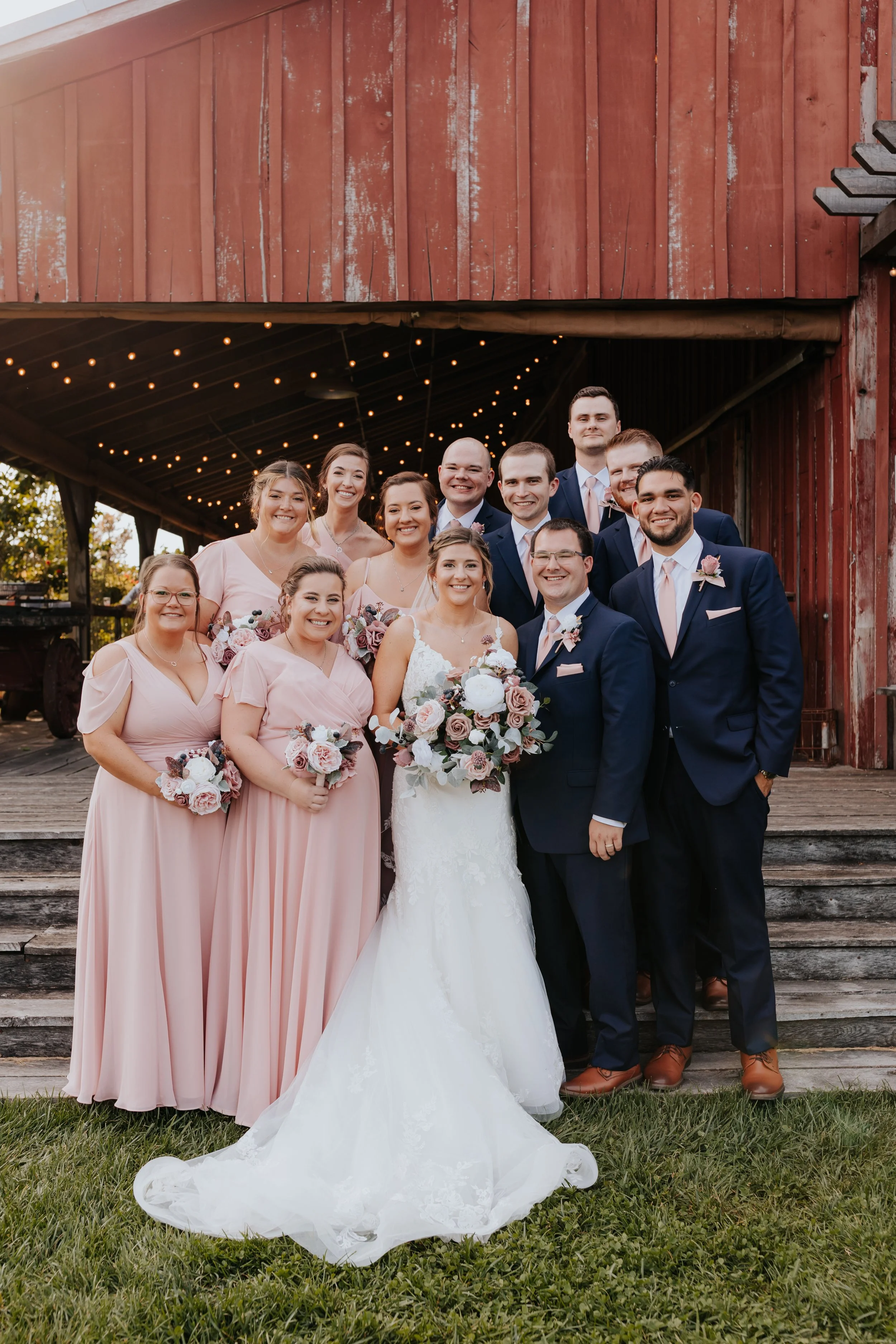 A wedding party of fifteen people, including the bride in a white wedding dress holding a bouquet, and the groom in a navy suit with a pink tie. The bridesmaids wear pink dresses, and the groomsmen wear navy suits with pink ties. They are standing ou