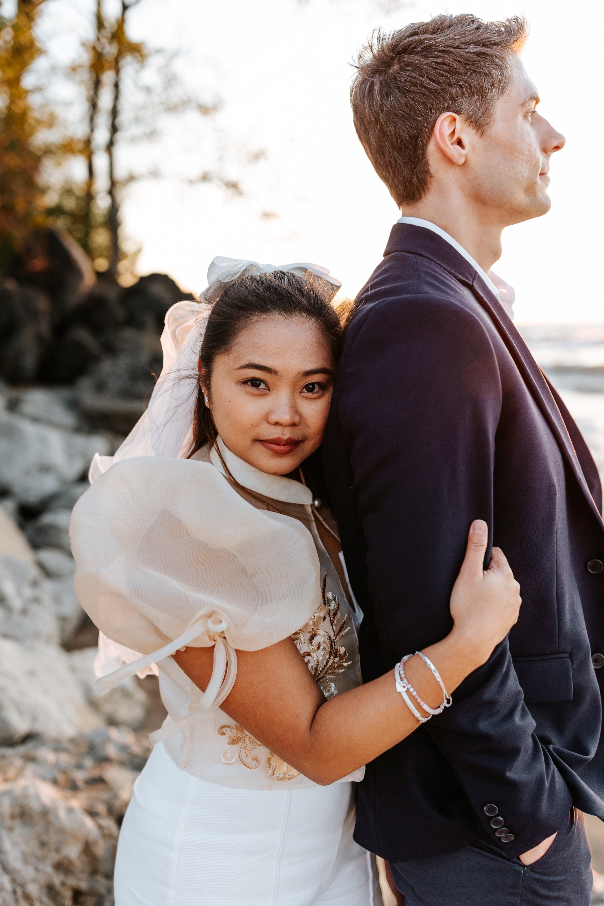 A woman in wedding attire hugging a man in a suit at the beach during sunset.
