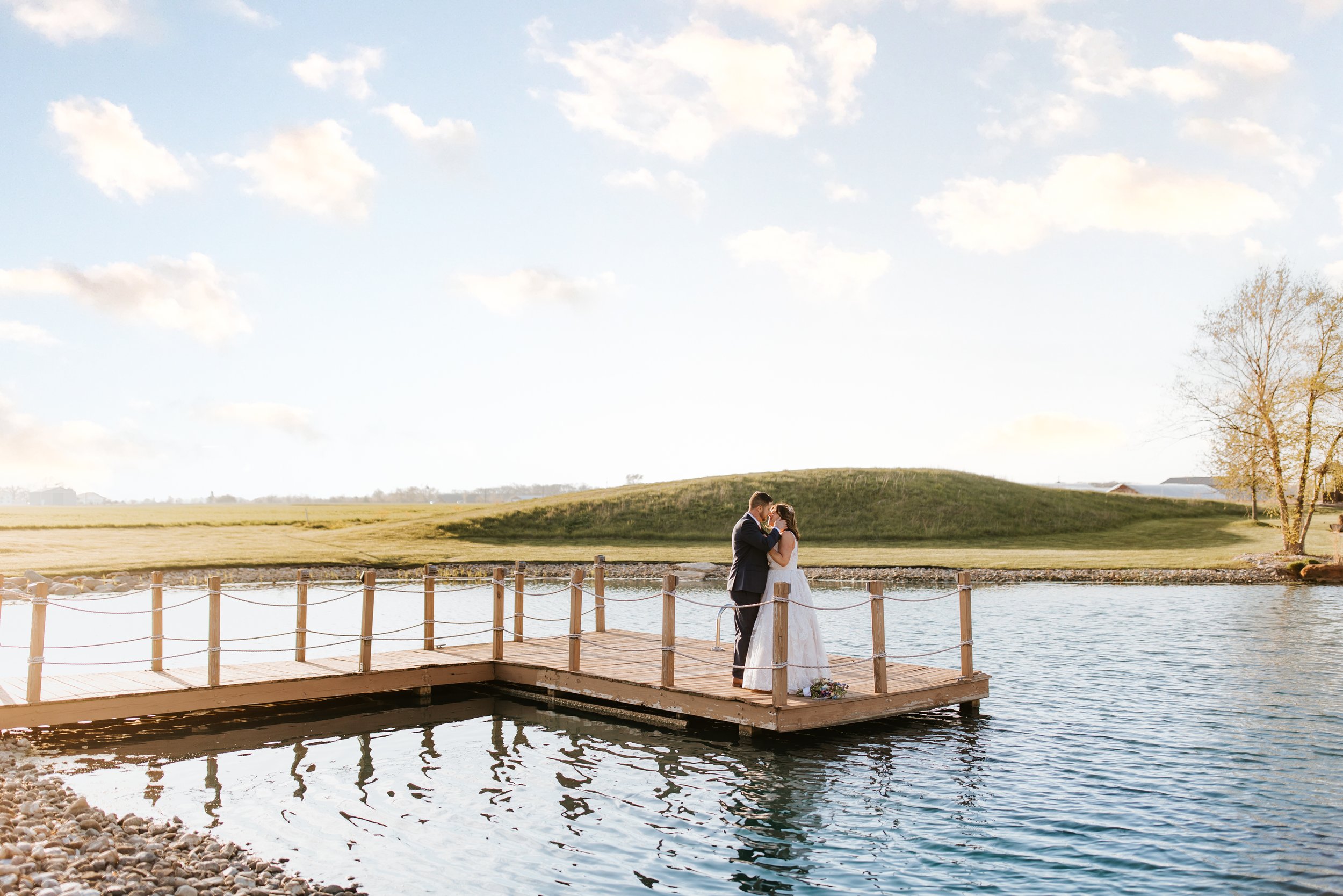 A bride and groom standing on a wooden dock by a lake, sharing a kiss during their wedding, with a grassy hill and trees in the background on a clear day.