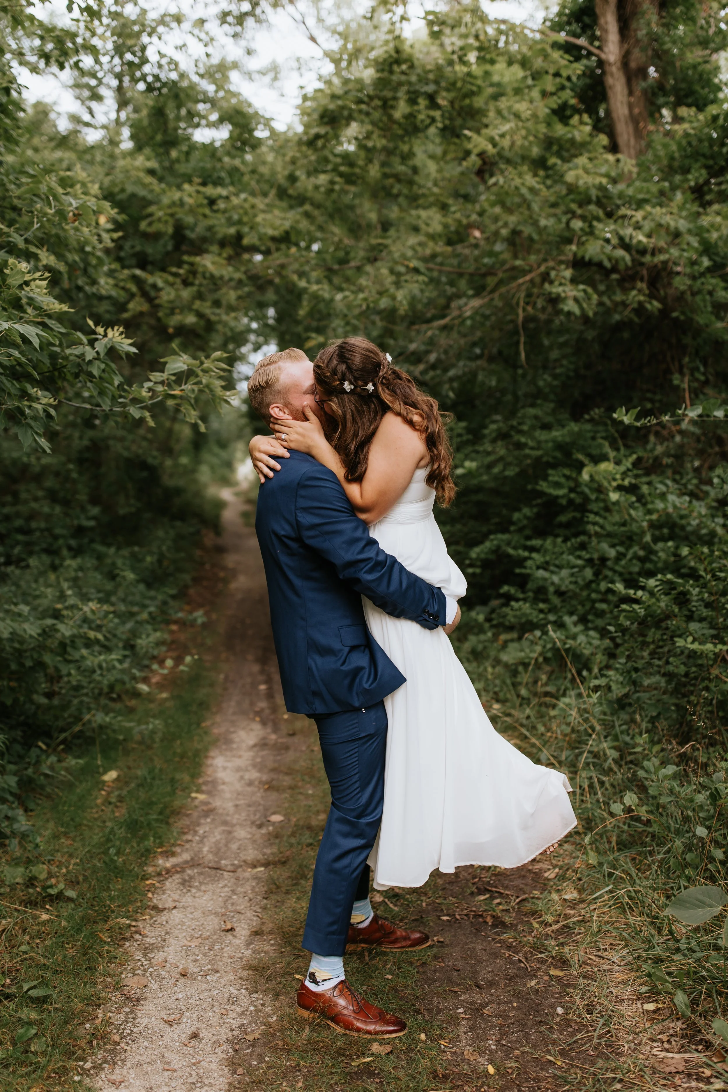A couple in wedding attire kissing in a wooded outdoor setting, with the man lifting the woman. The woman has long brown hair with a floral headpiece, and the man has blonde hair. The woman is wearing a white dress, and the man is in a blue suit.