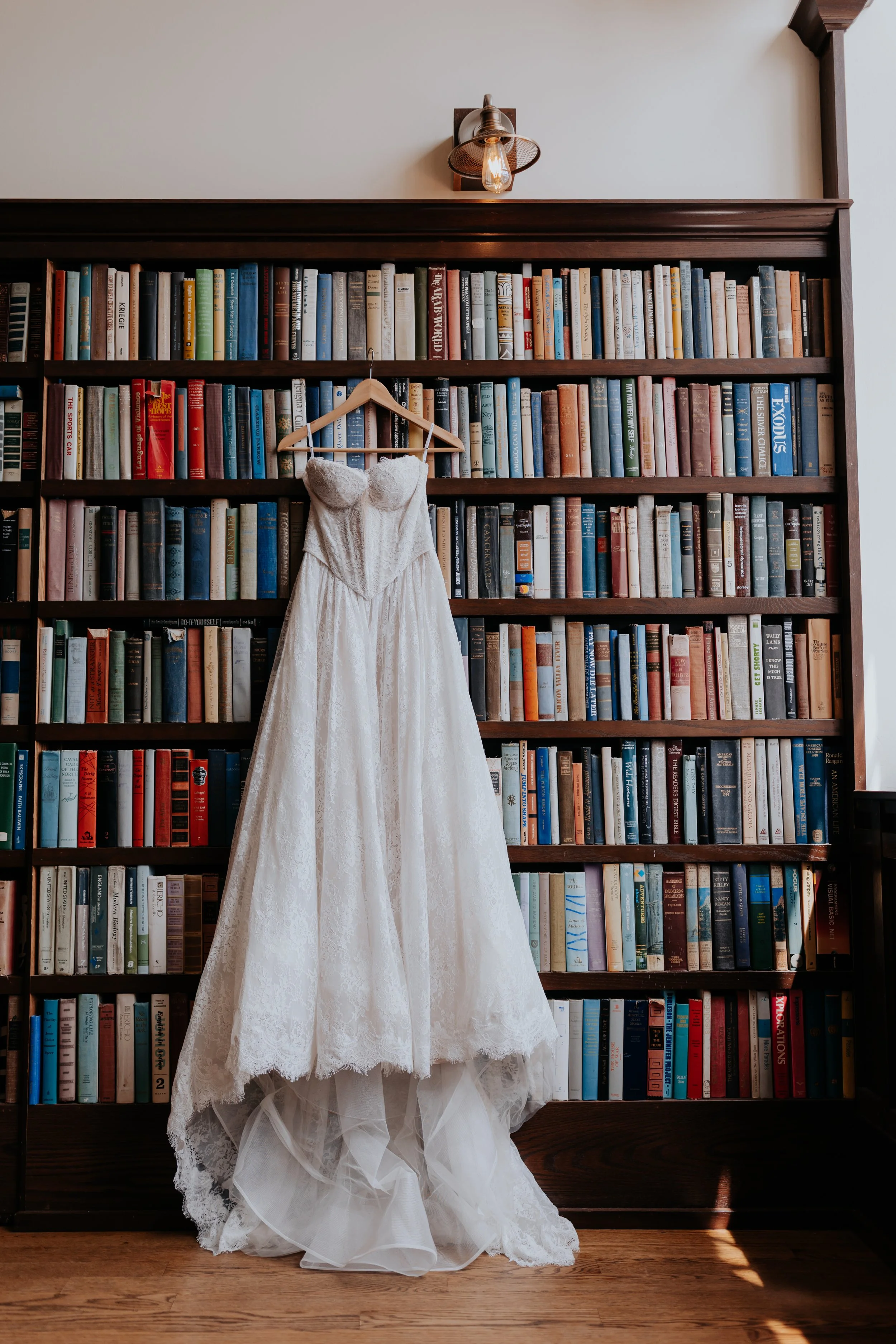 Wedding dress hanging on a wooden hanger against a bookshelf filled with books in a room with wooden floors.