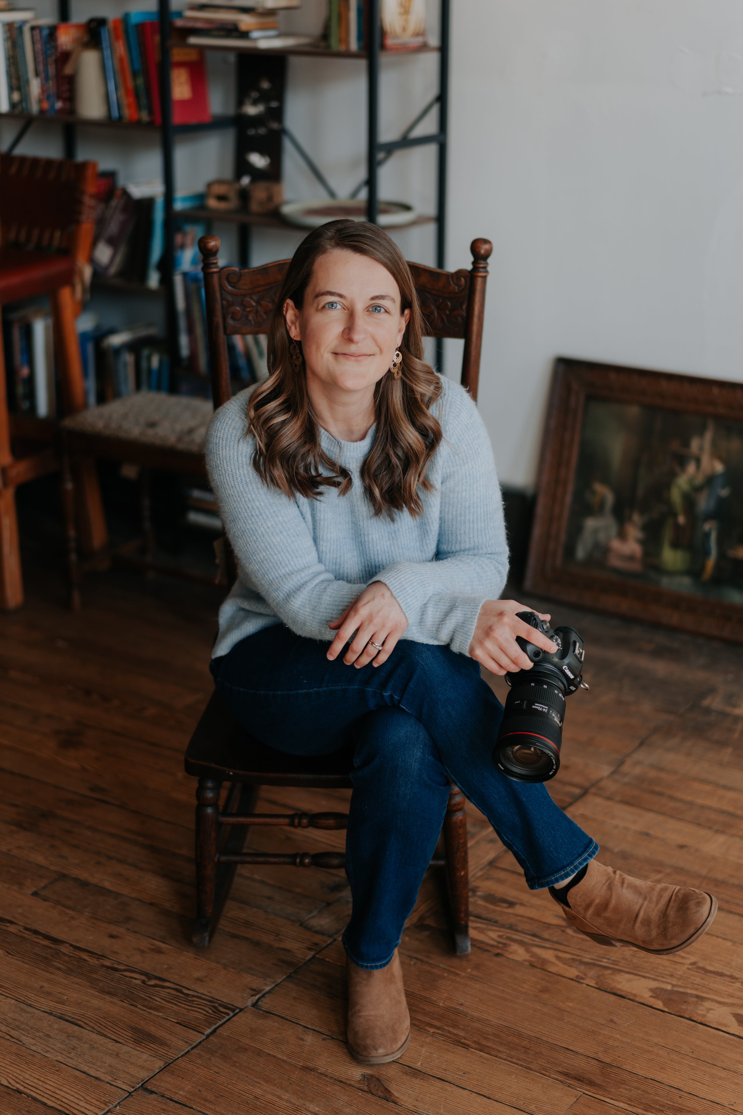 A woman with brown wavy hair, wearing a light gray sweater, blue jeans, and brown boots, sitting cross-legged on a wooden chair, holding a professional camera, in a room with bookshelves and a framed painting on the floor.