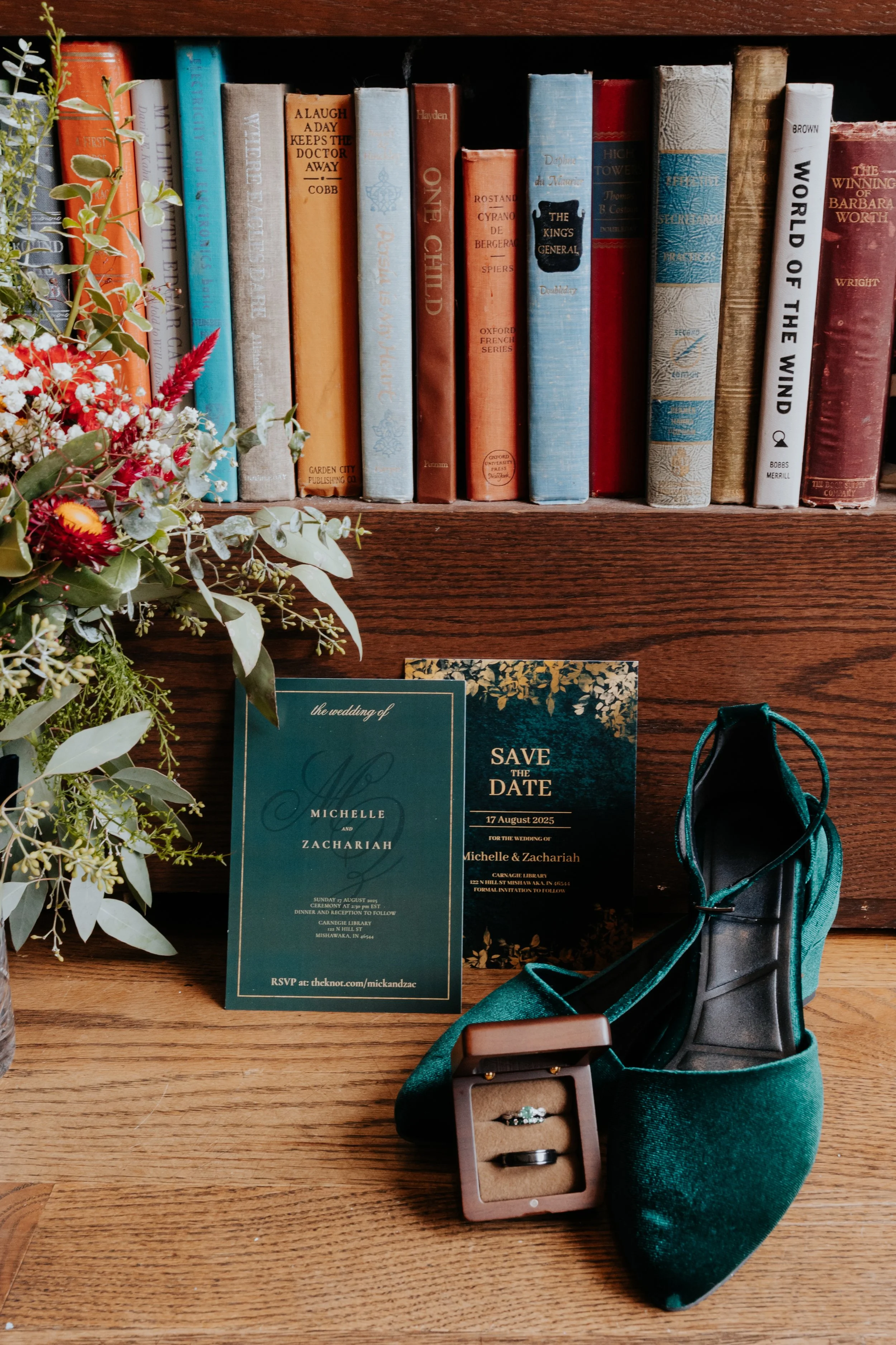 Wedding invitation, green dance shoe, engagement ring in a box, and a bouquet with greenery and red flowers on a wooden surface in front of a bookshelf.