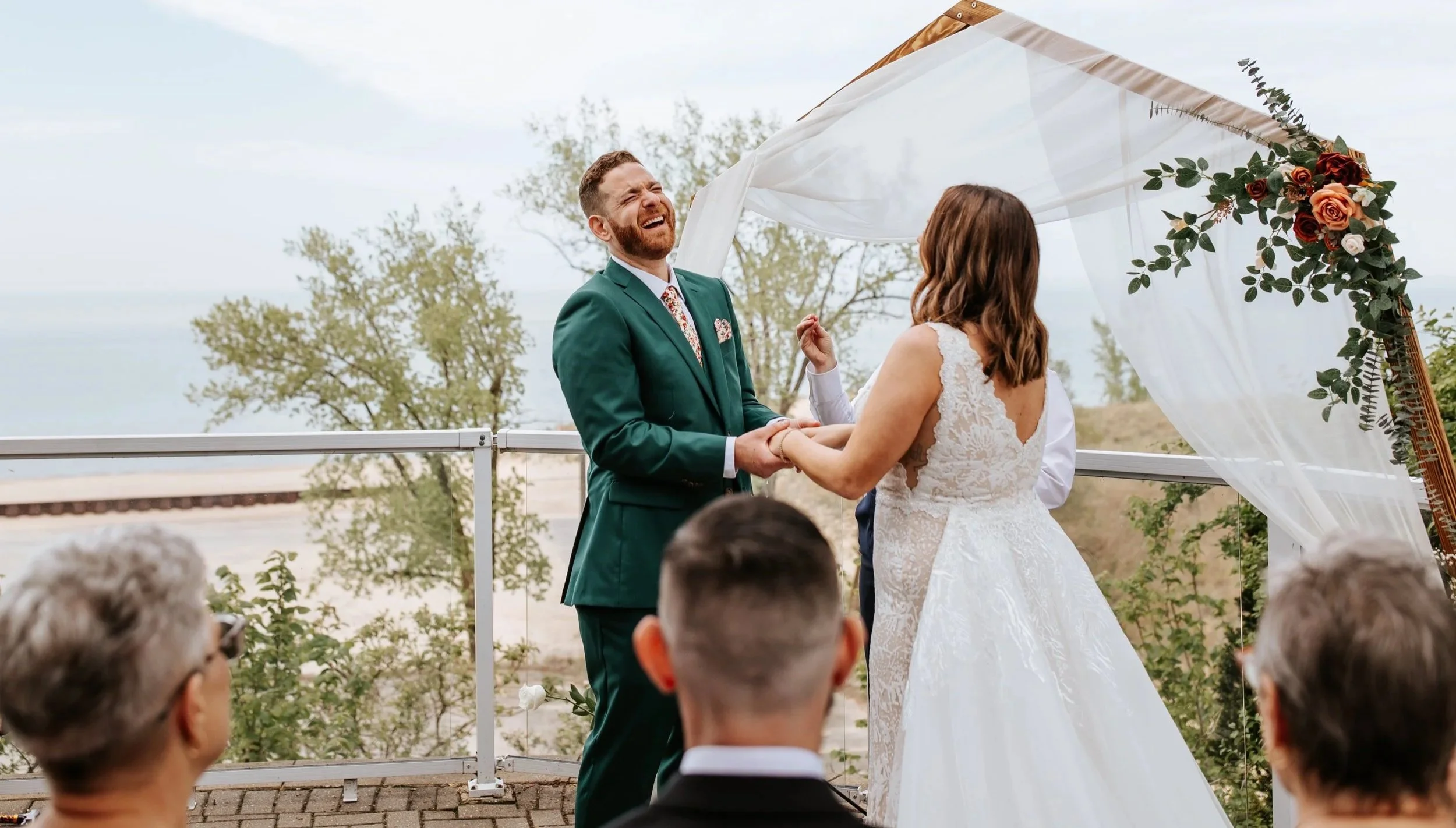 A wedding ceremony taking place outdoors with a couple holding hands, exchanging vows under a floral arch with draped white fabric, overlooking a scenic landscape with trees and water in the background. Guests are seated nearby.