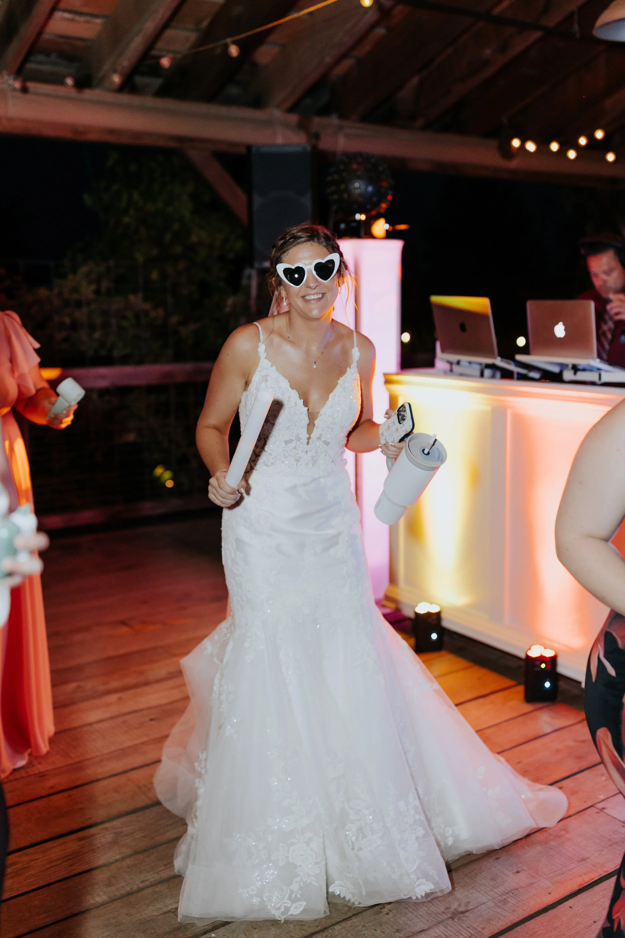 A woman in a wedding dress wearing heart-shaped sunglasses at a wedding reception, holding a glass and a cloth, smiling.