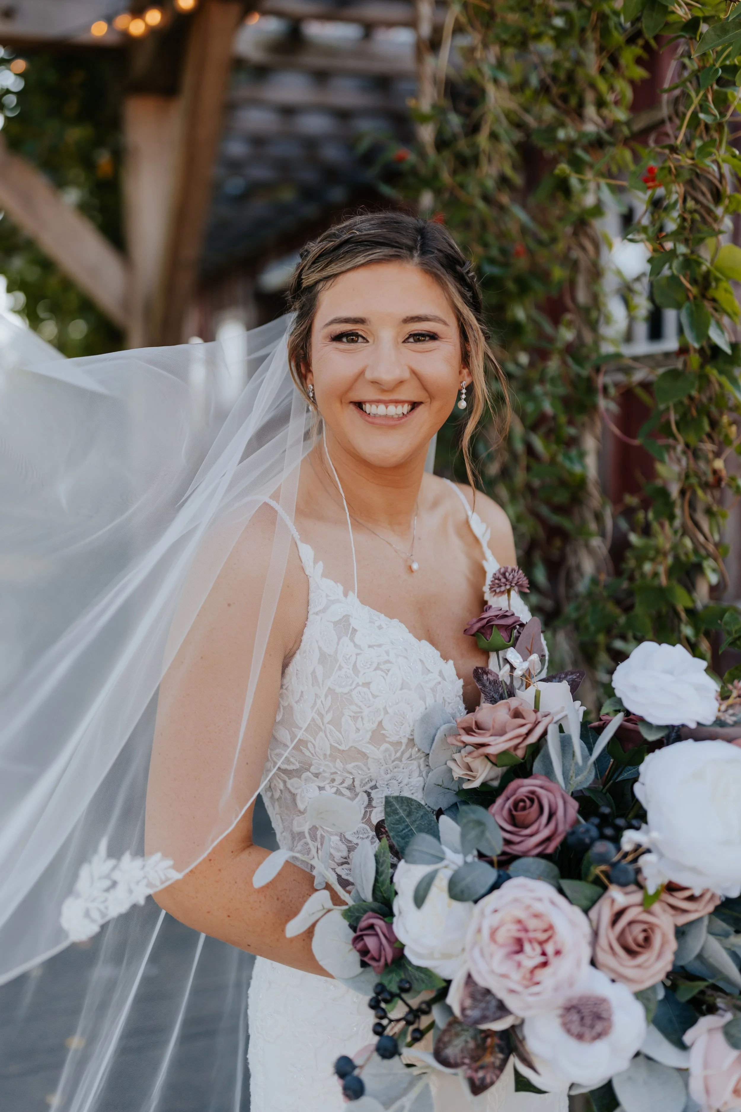 A smiling bride holding a bouquet of roses, peonies, and greenery, outdoors with a wooden structure and vines in the background.