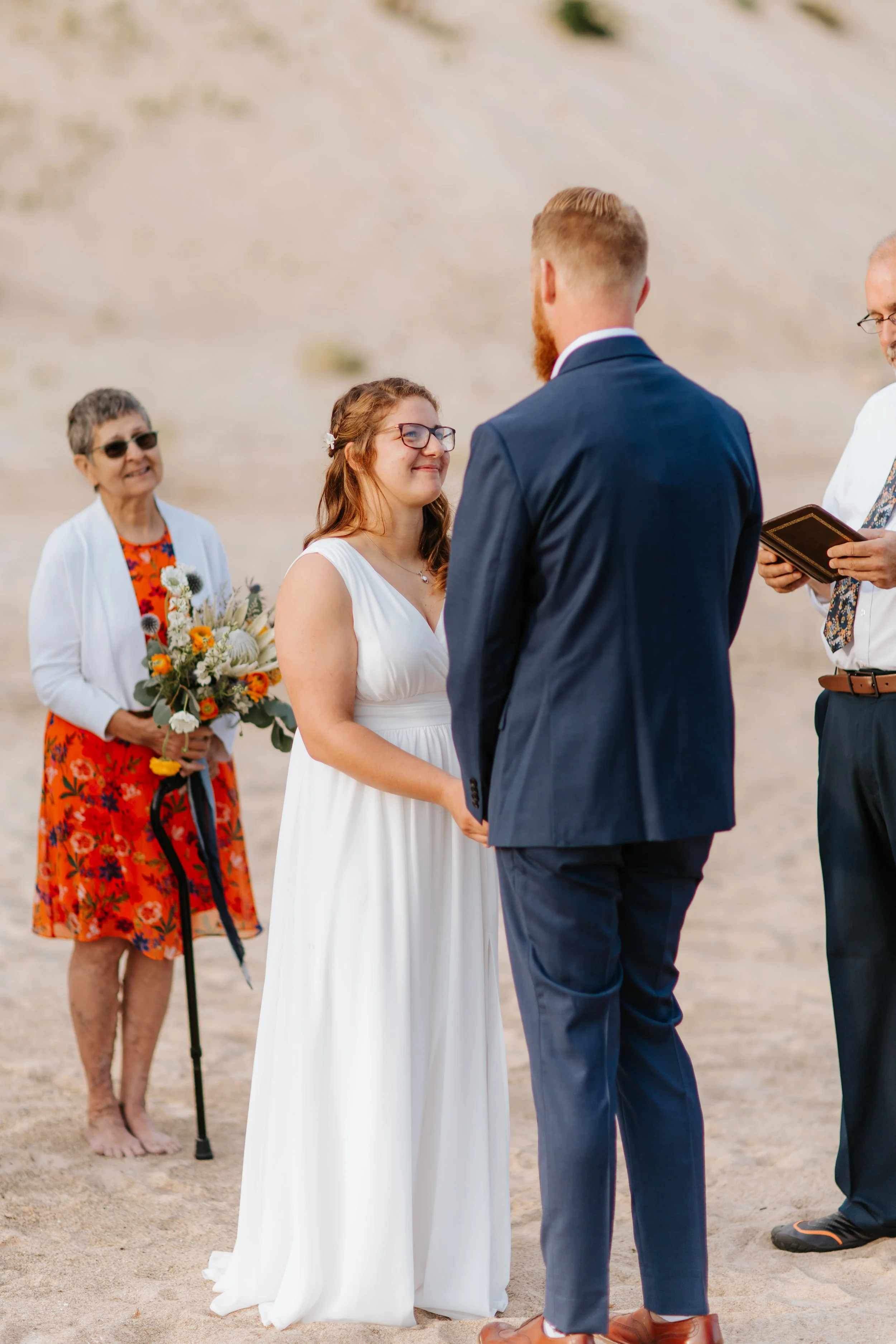 A couple getting married on the beach, holding hands, with an officiant reading vows. Two women, one holding a bouquet, watch the ceremony.