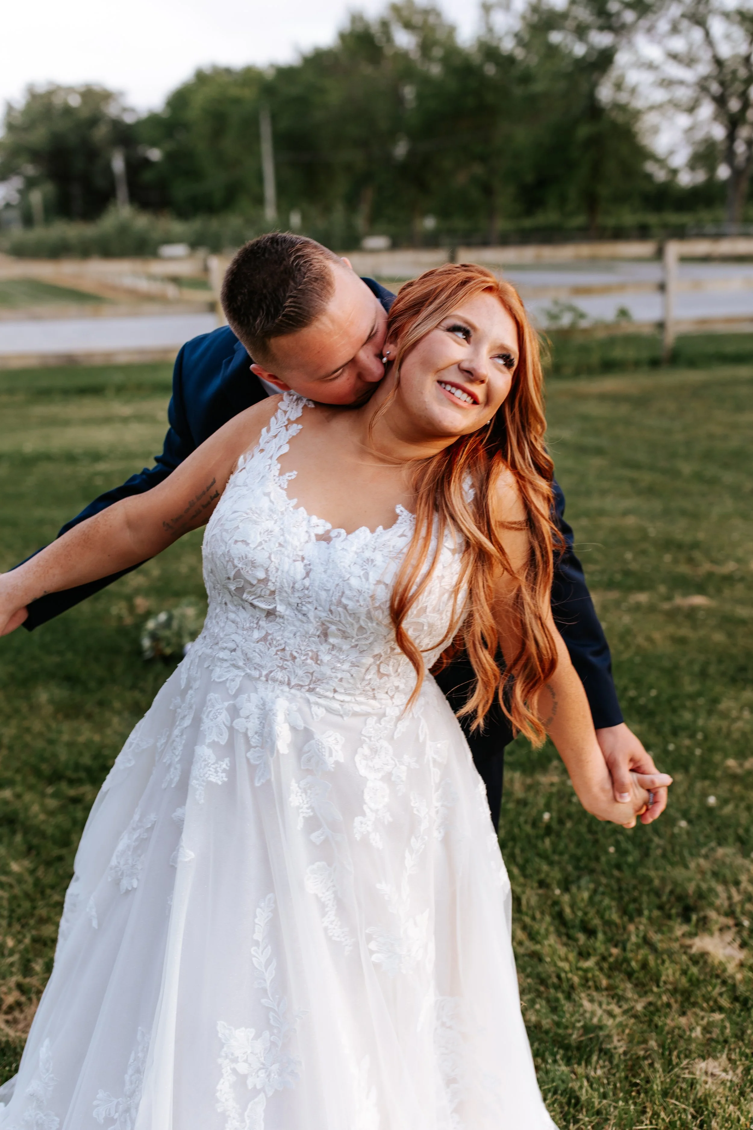 A bride with long red hair wearing a white lace wedding dress, and a groom in a dark suit are holding hands in a grassy outdoor setting. The groom is leaning in to kiss the bride's neck, and she is smiling.