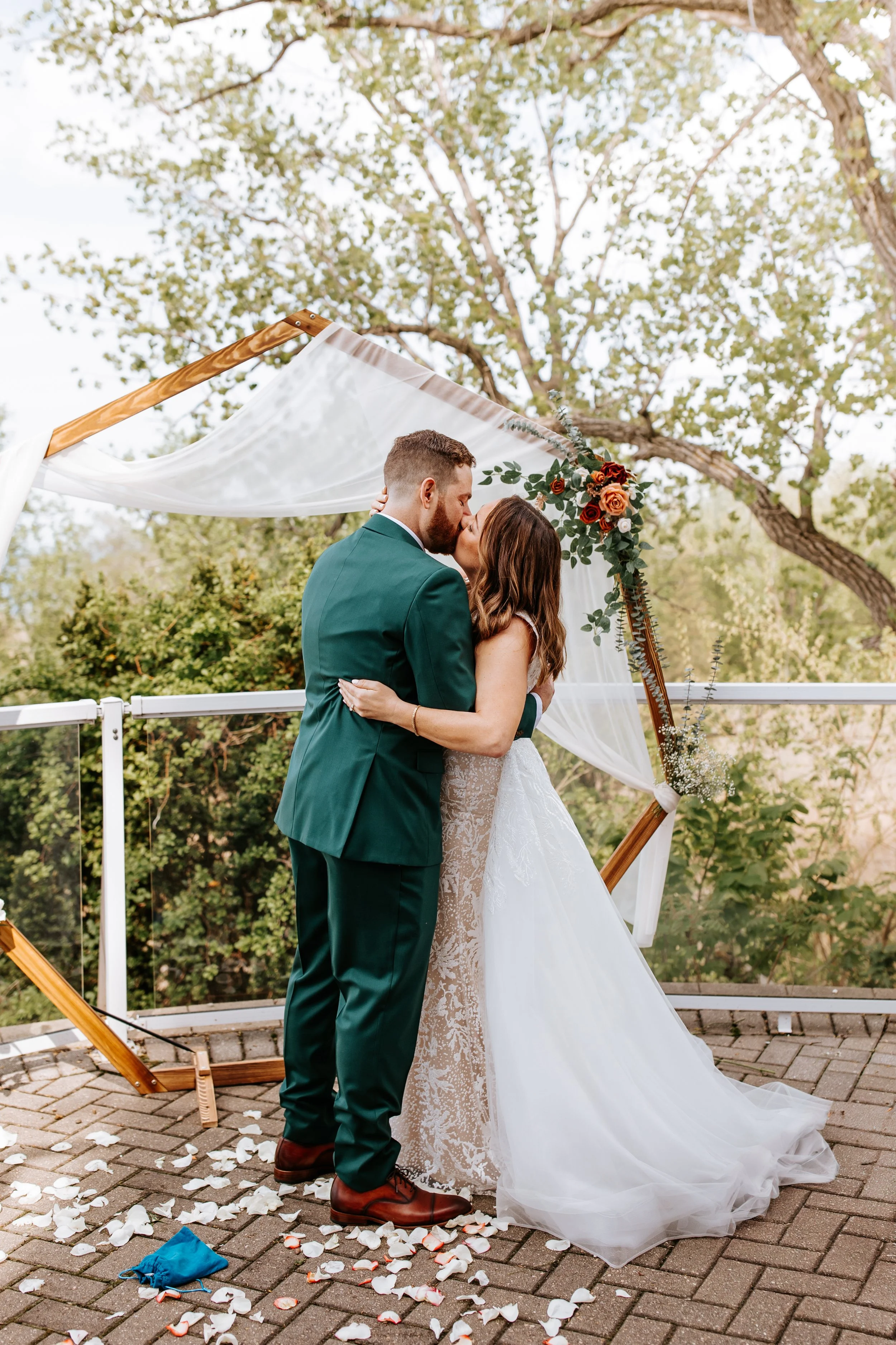 A bride and groom share a kiss during their outdoor wedding ceremony, standing under a wooden arch decorated with white drapery and red and orange flowers, on a brick terrace with rose petals scattered on the ground.