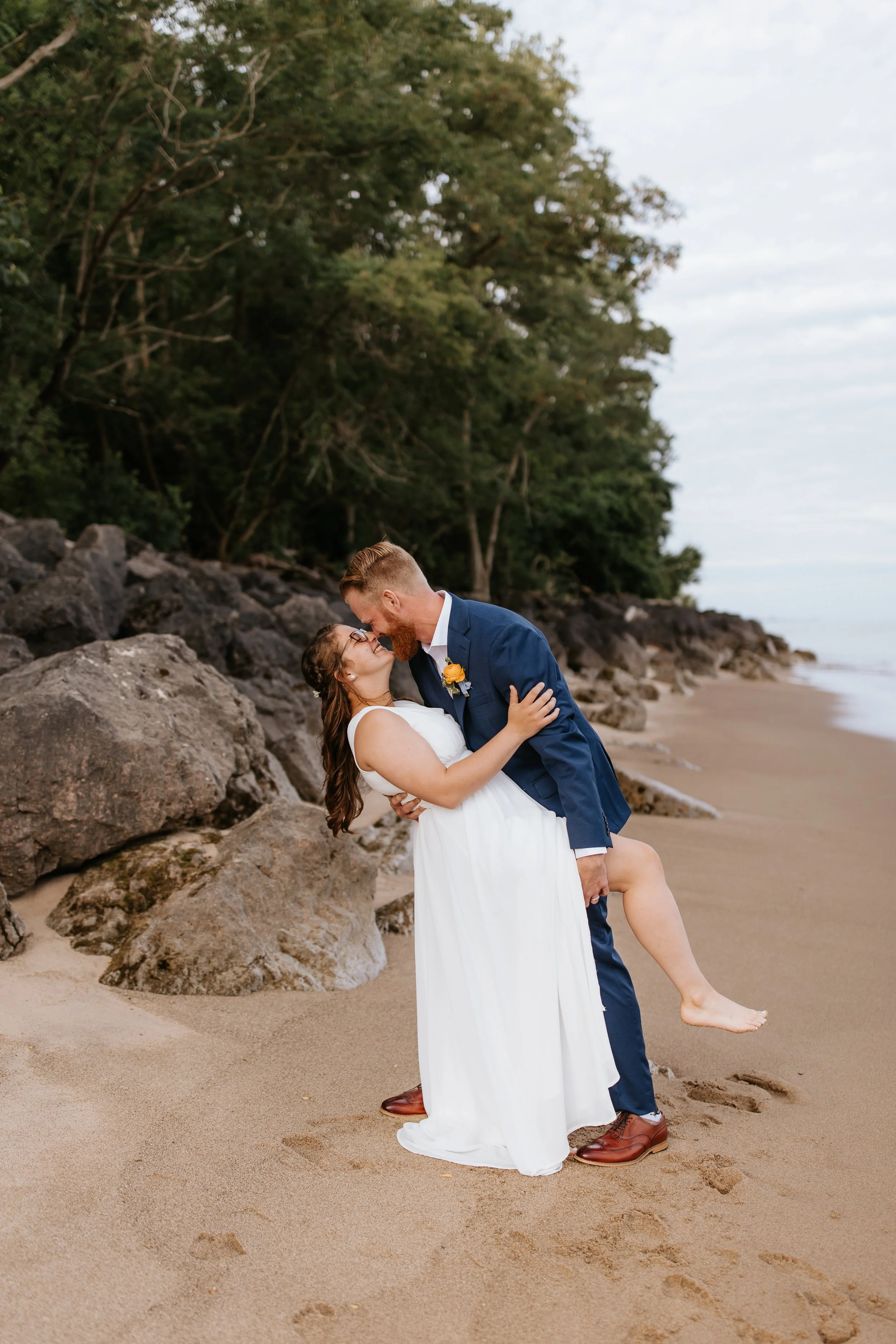 A couple in wedding attire standing on a sandy beach with rocky formations and greenery in the background, sharing a close moment as he dips her.