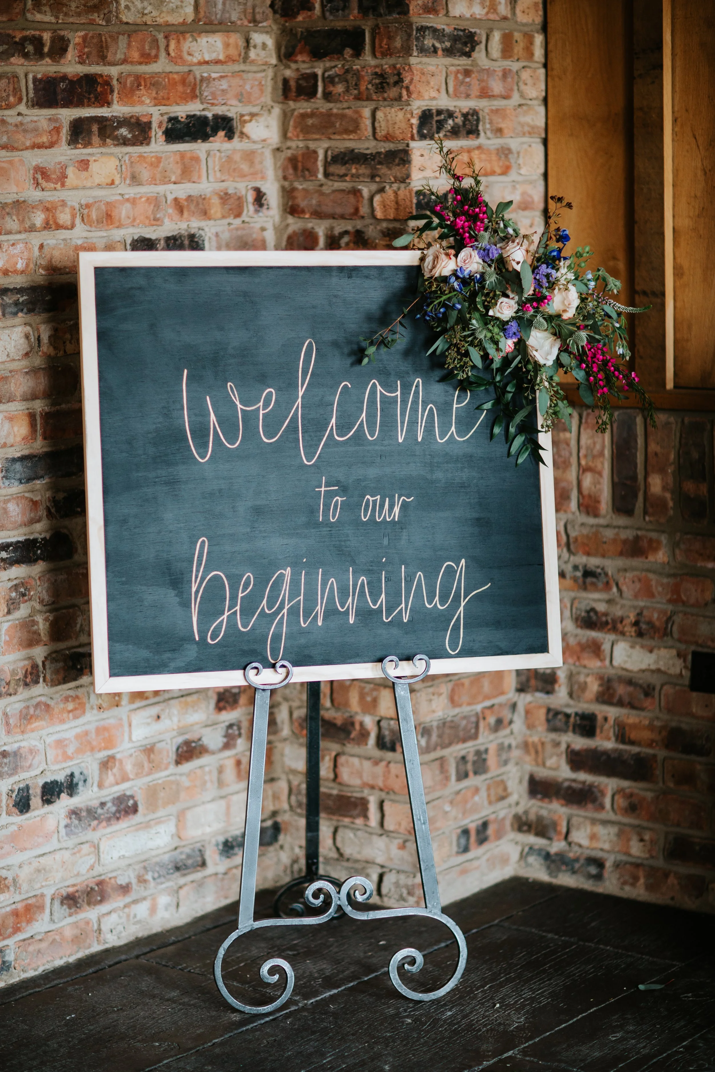 A welcome sign on an easel with a dark green background and a floral arrangement at the top right corner, placed against a brick wall.