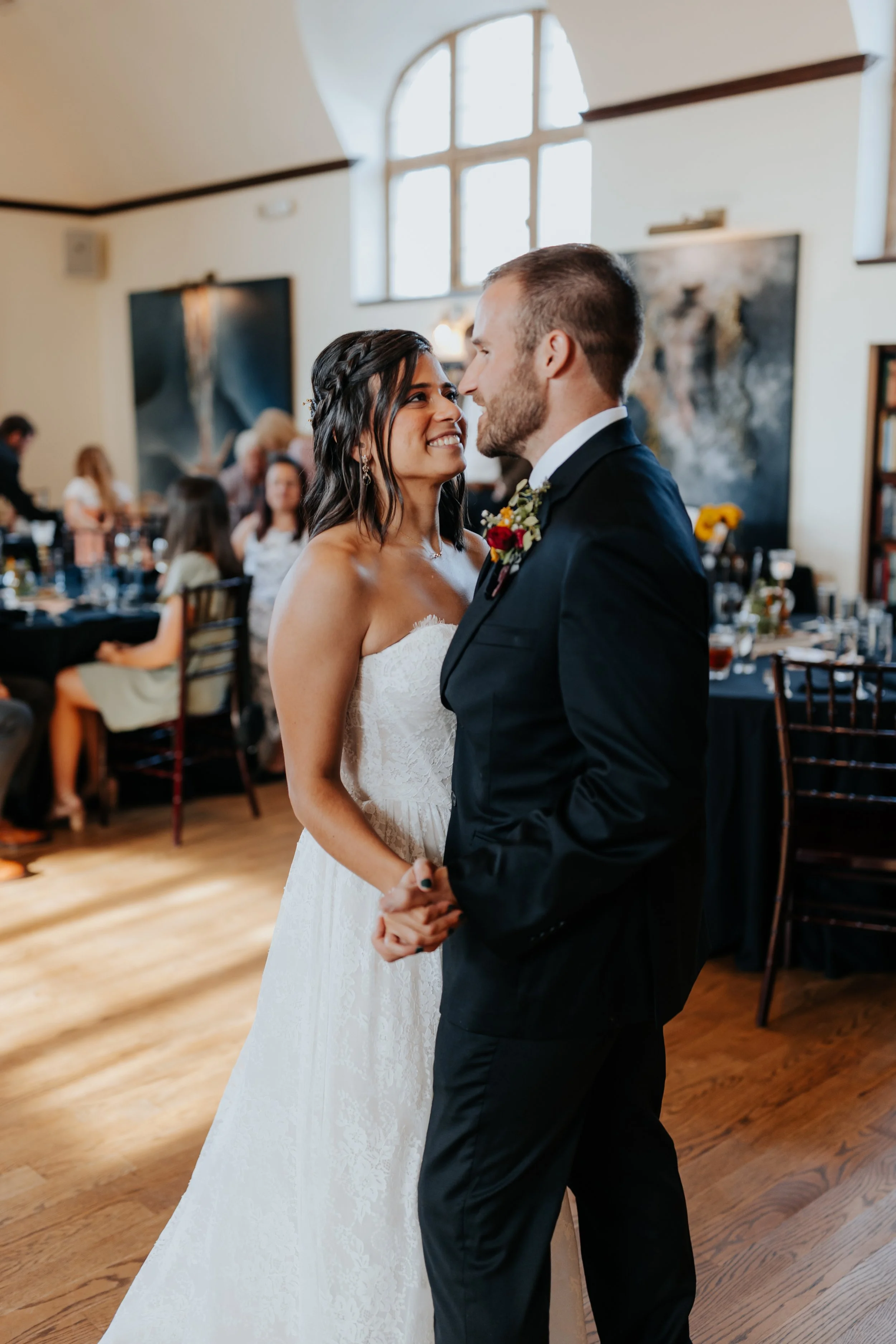 Bride and groom holding hands during their wedding dance in a decorated venue, smiling at each other.