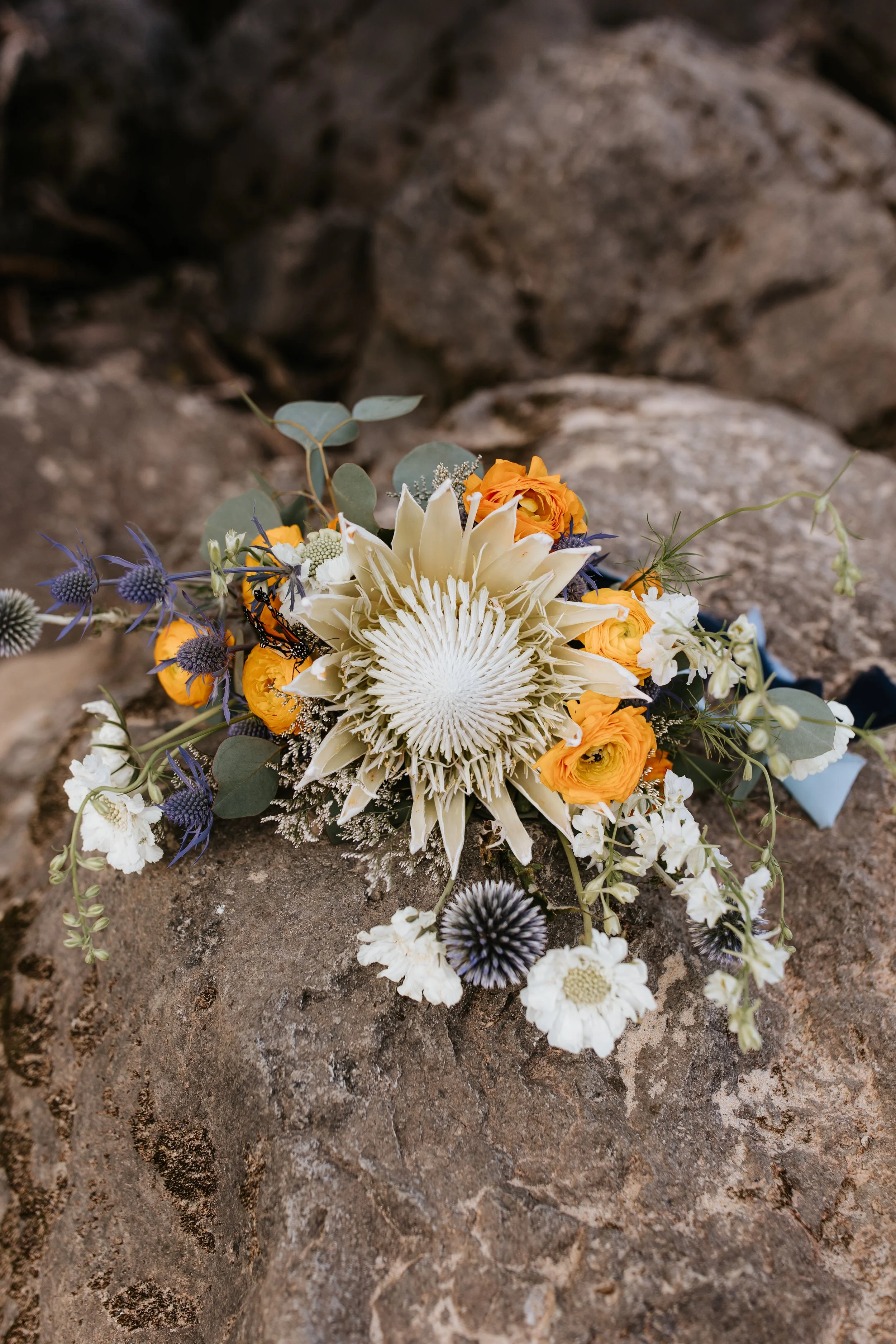A vibrant floral arrangement with orange roses, white and purple flowers, and greenery placed on a rocky surface.