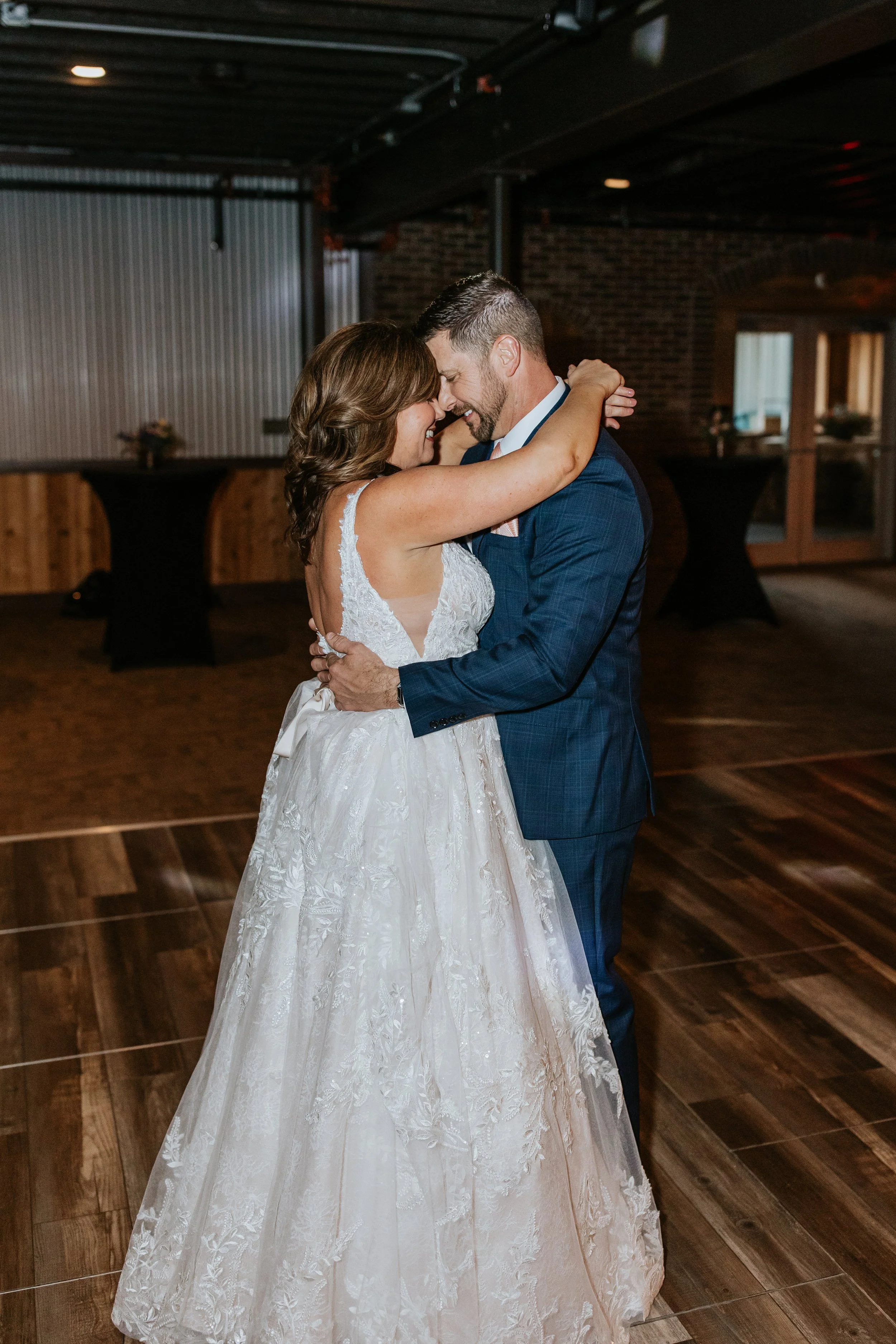 A bride in a white lace wedding dress and a groom in a dark suit dancing closely in a rustic indoor wedding venue.