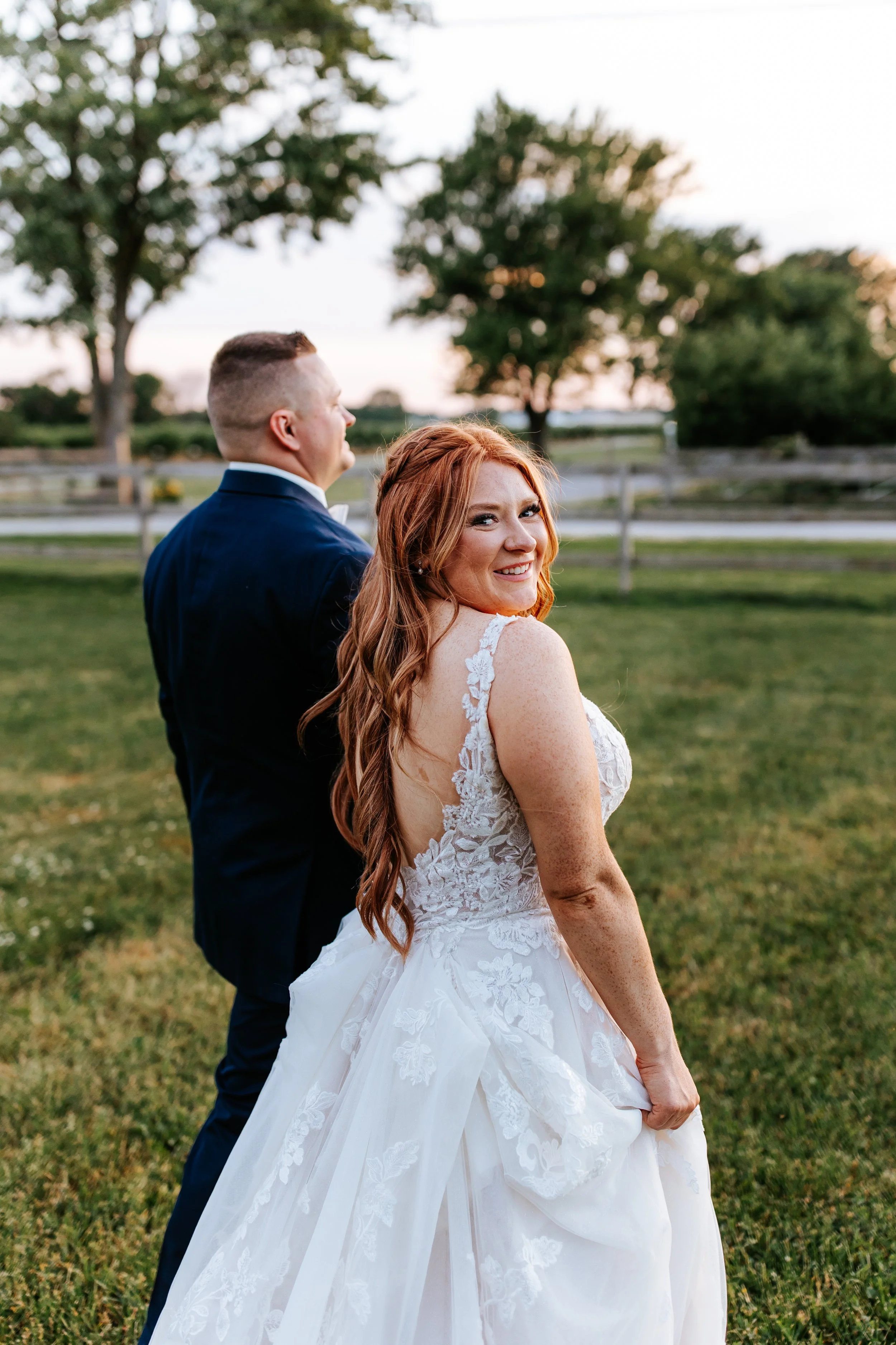 A smiling bride with red hair in a white wedding dress holds her dress and stands in a grassy field with trees and a wooden fence in the background at sunset, with the groom in a dark suit behind her.