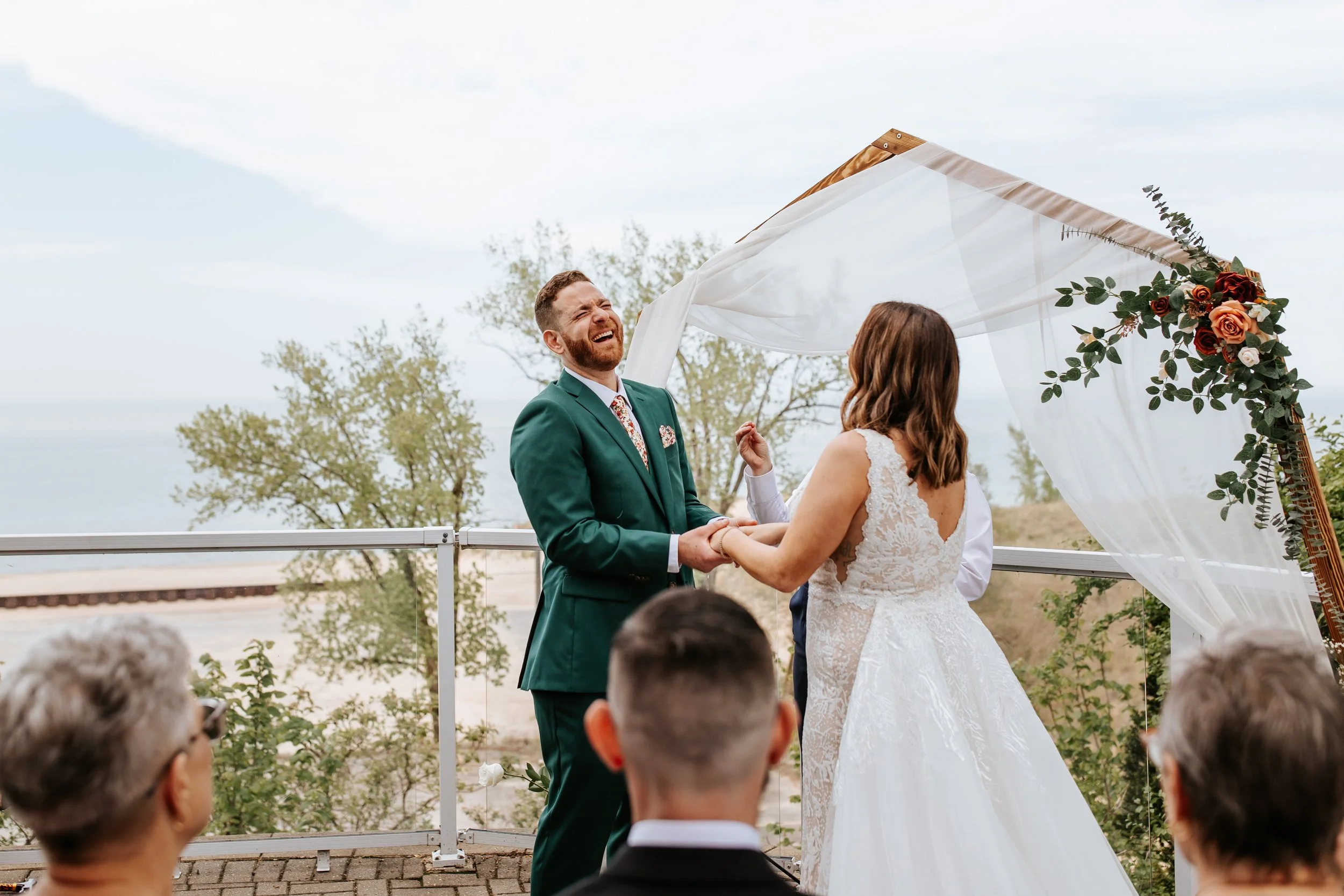 A couple getting married outdoors under a decorated arch, with the groom in a green suit laughing and holding hands with the bride in a white lace wedding dress, surrounded by guests.
