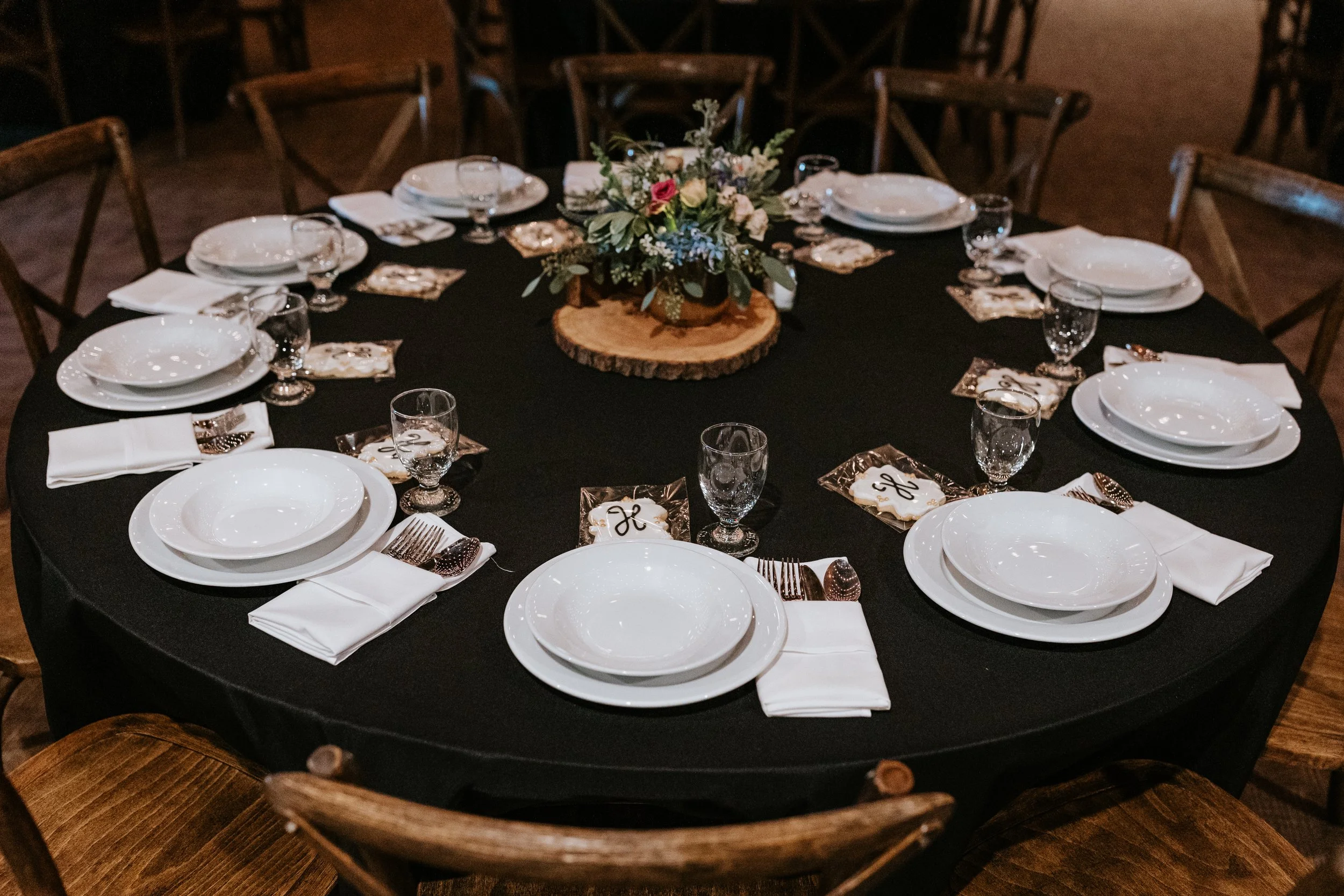 Round table set for a formal event, with a black tablecloth, white plates, silverware, wine glasses, white napkins, and a floral centerpiece on a wooden slab.