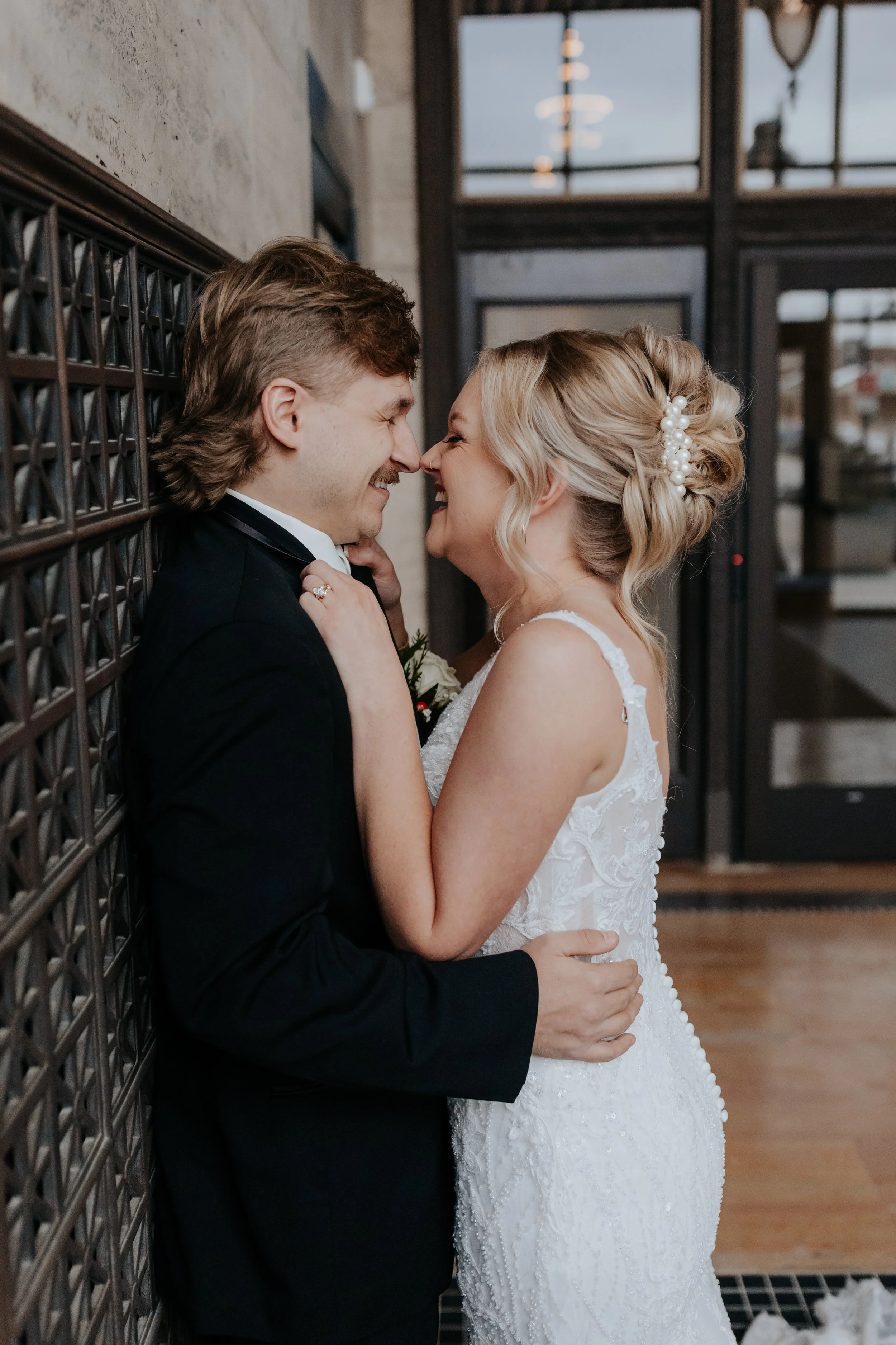A newlywed couple is smiling and leaning in close, touching their noses together inside a building. The bride is in a white lace wedding dress with her hair up decorated with pearls, and the groom is in a black suit. They are standing on a wooden floor beside a wall with a decorative metal grid.