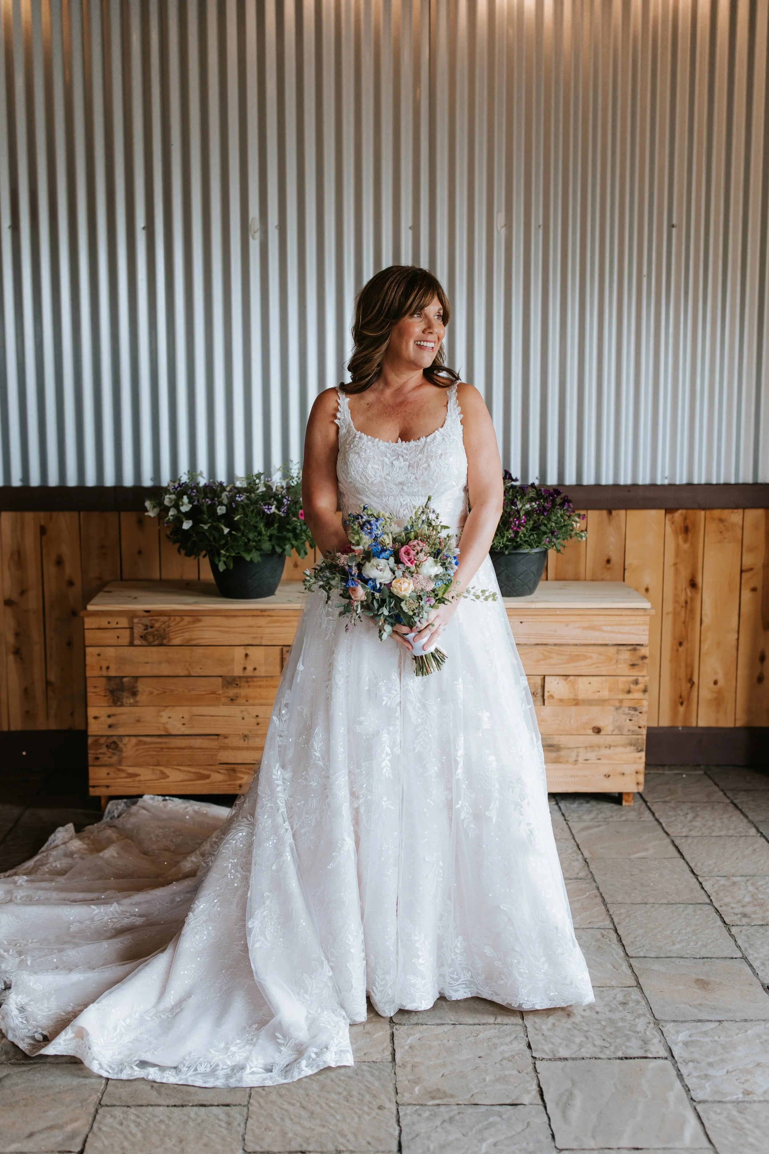 A bride in a white lace wedding dress holding a bouquet of flowers, standing indoors in front of a wooden and metal backdrop with potted plants.