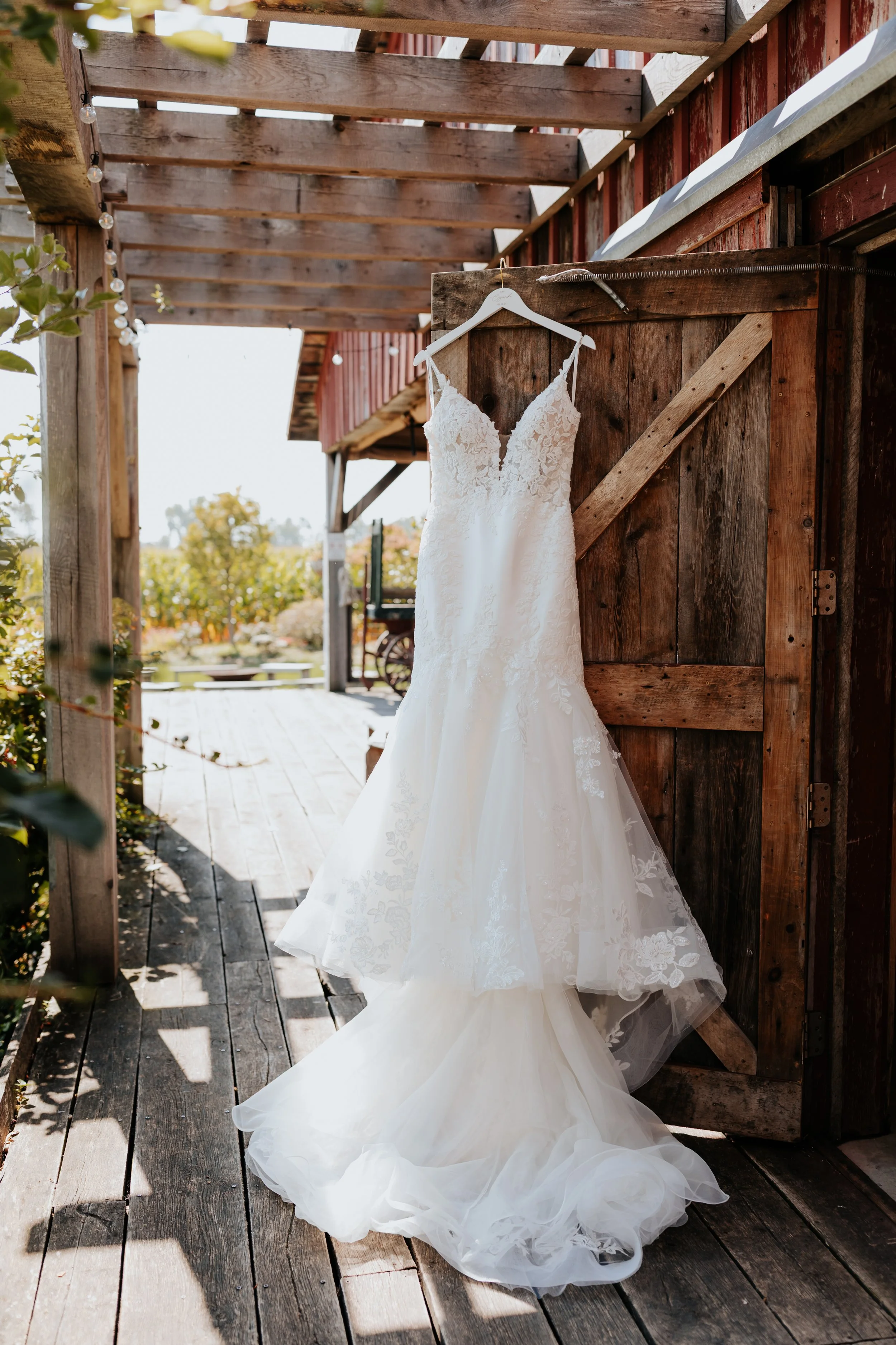 A white wedding dress hanging on a white hanger outside on a wooden barn door with rustic surroundings.