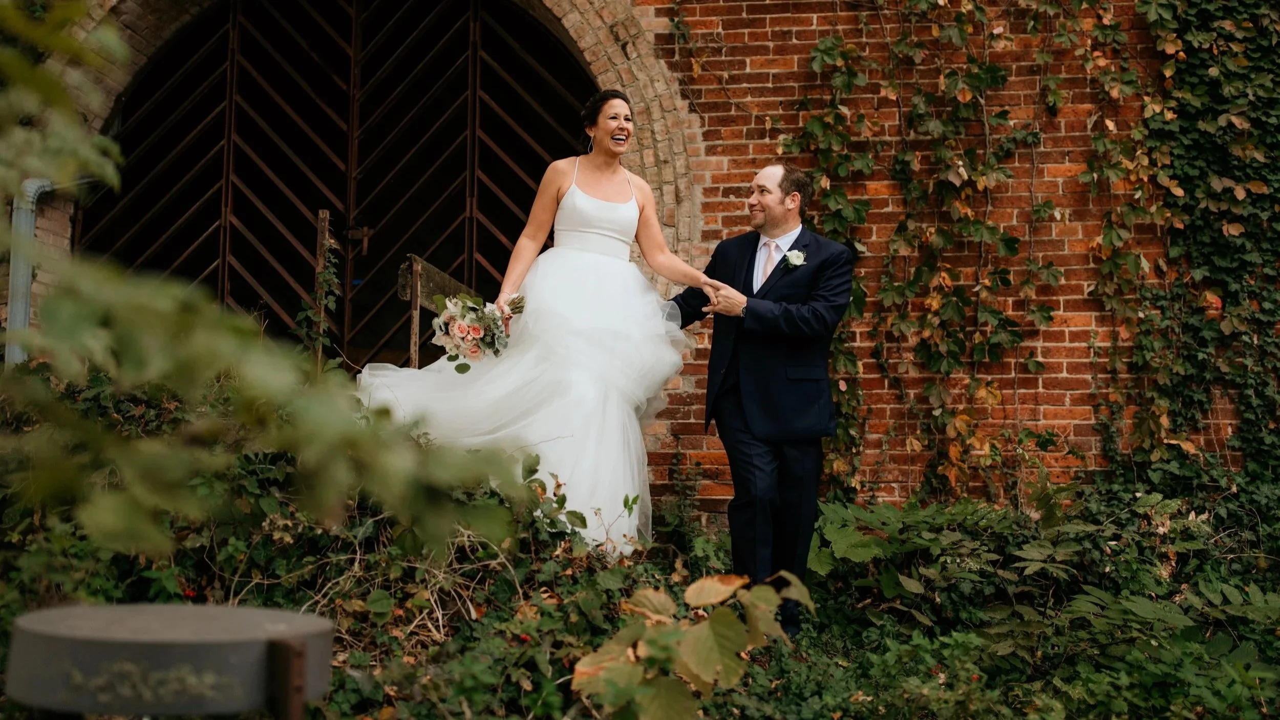 A bride in a white wedding dress holding a bouquet and a groom in a dark suit with a white boutonniere, standing outdoors in front of a brick wall covered with green ivy, smiling and holding hands.