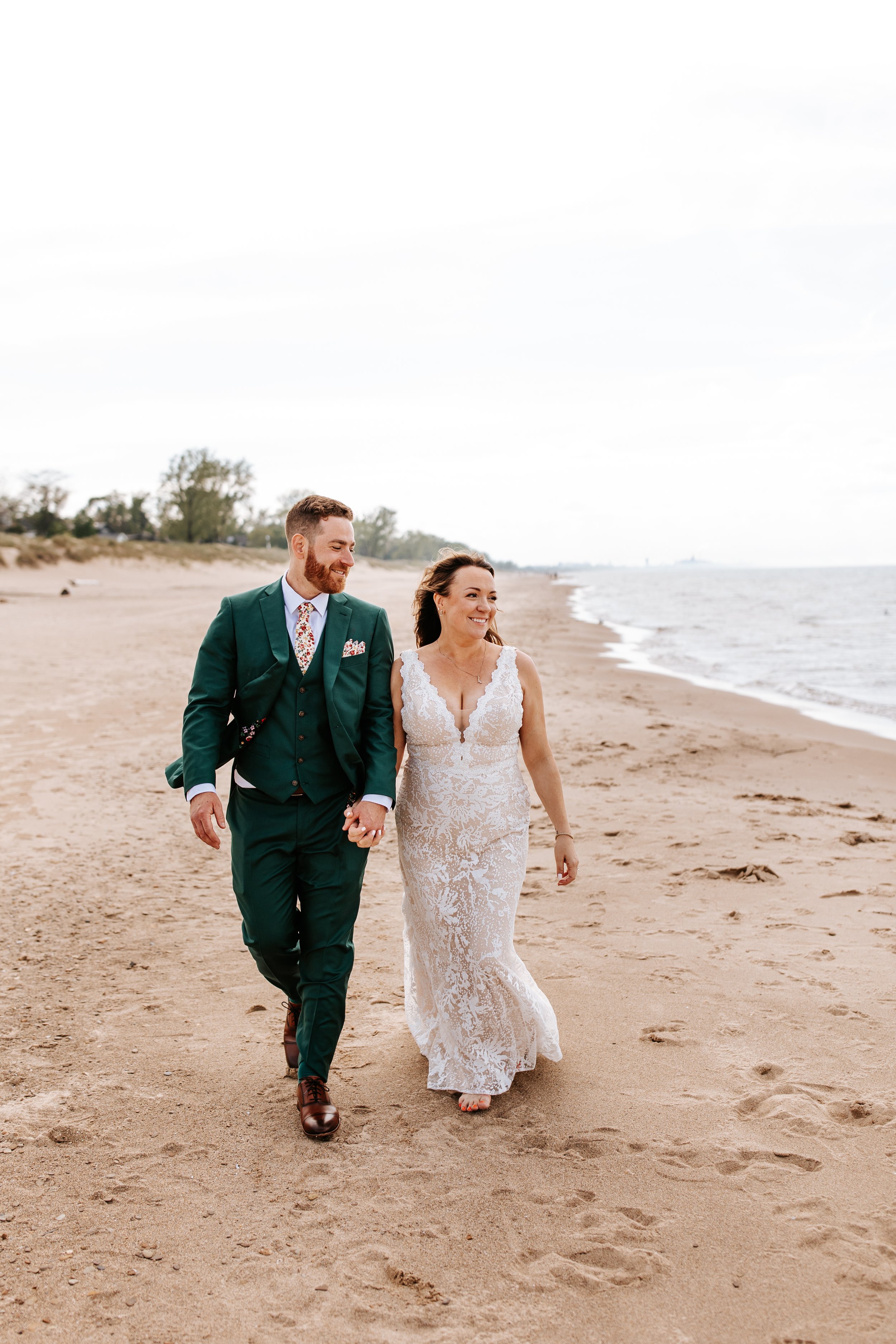 A smiling newlywed couple walking hand-in-hand on a sandy beach, dressed in wedding attire, with the ocean and a cloudy sky in the background.