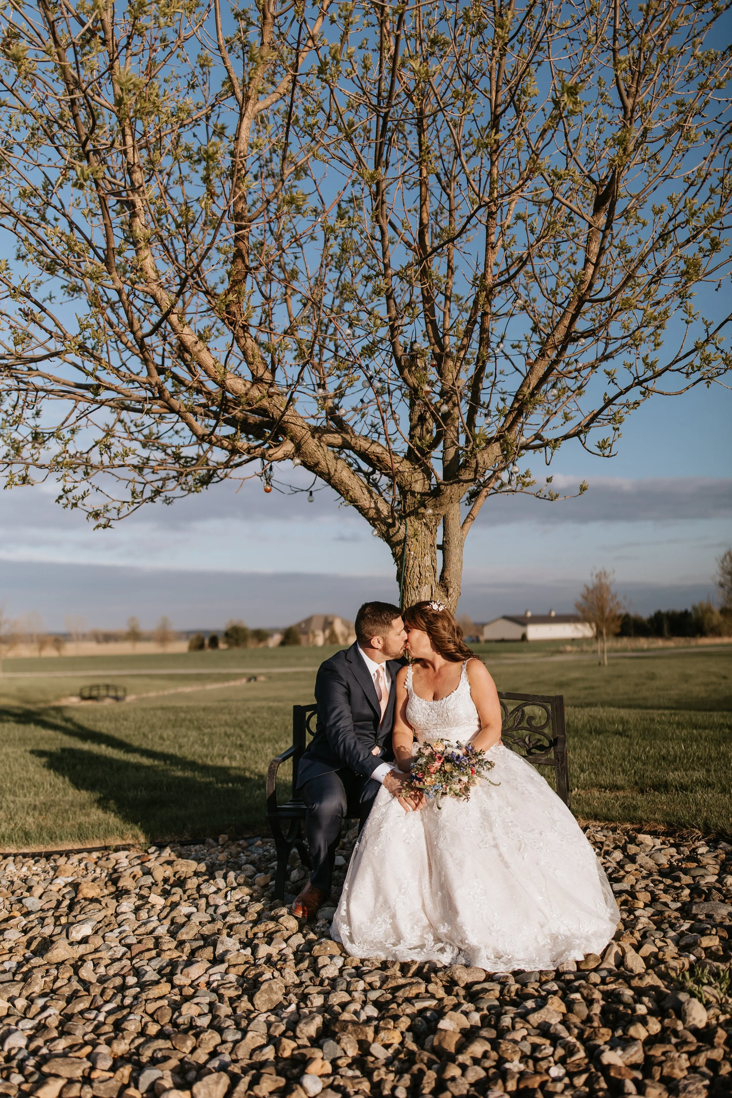 A bride and groom sitting on a bench beneath a large tree, holding hands and sharing a kiss in an outdoor setting with grass and houses in the background.