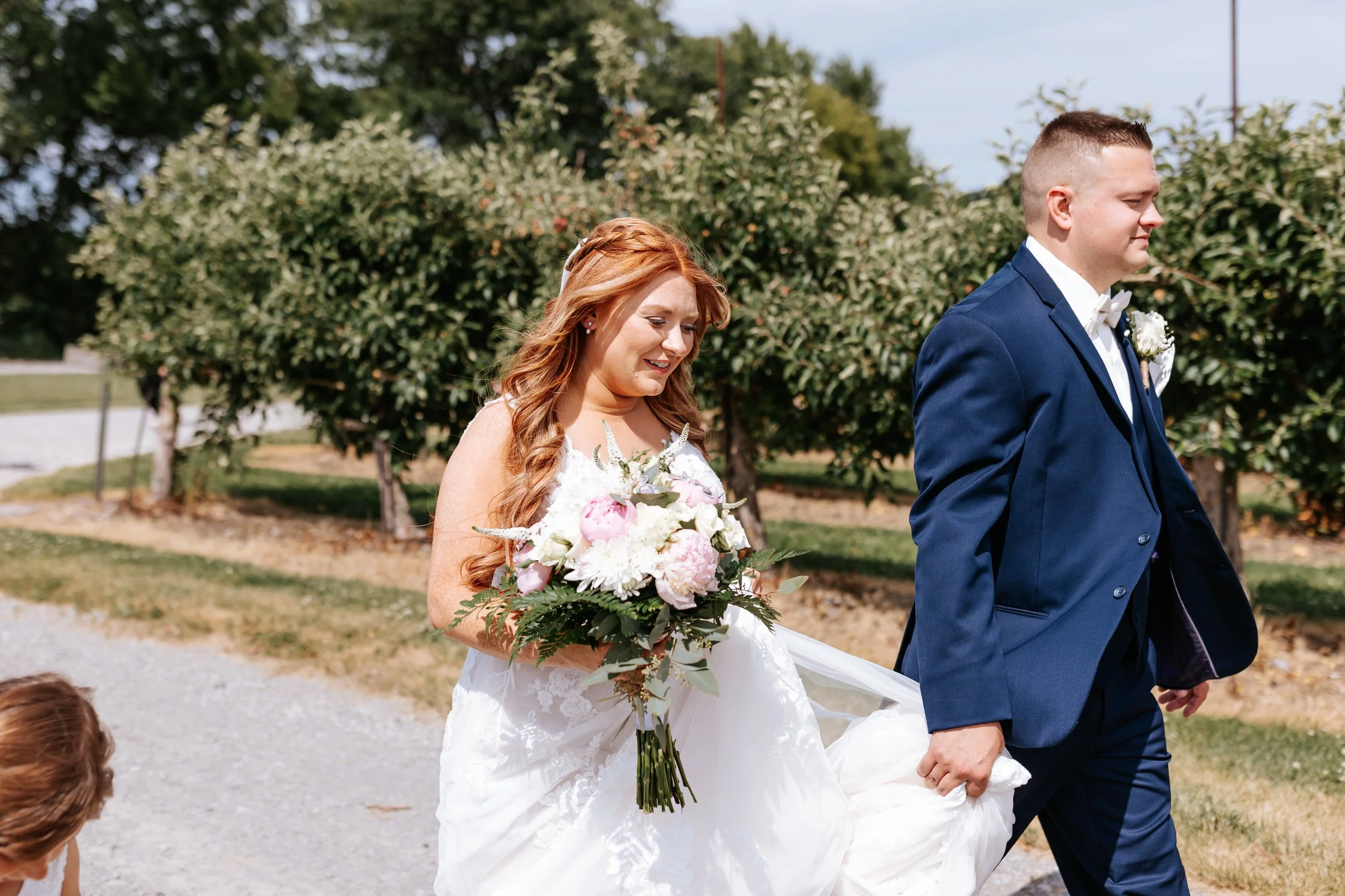 A bride and groom walking outdoors, the bride holding a bouquet of flowers and the groom holding her dress, during a wedding ceremony or photo session.