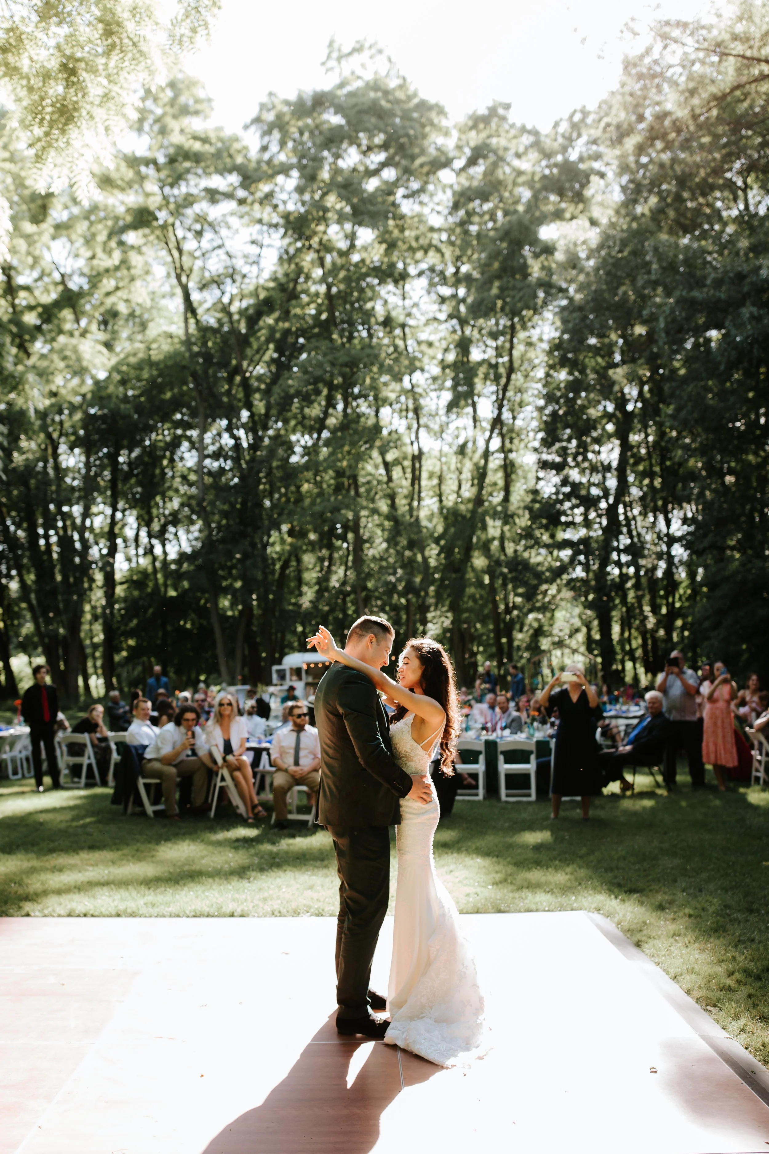 A bride and groom dancing during their wedding reception outdoors with guests seated at tables under trees in the background.