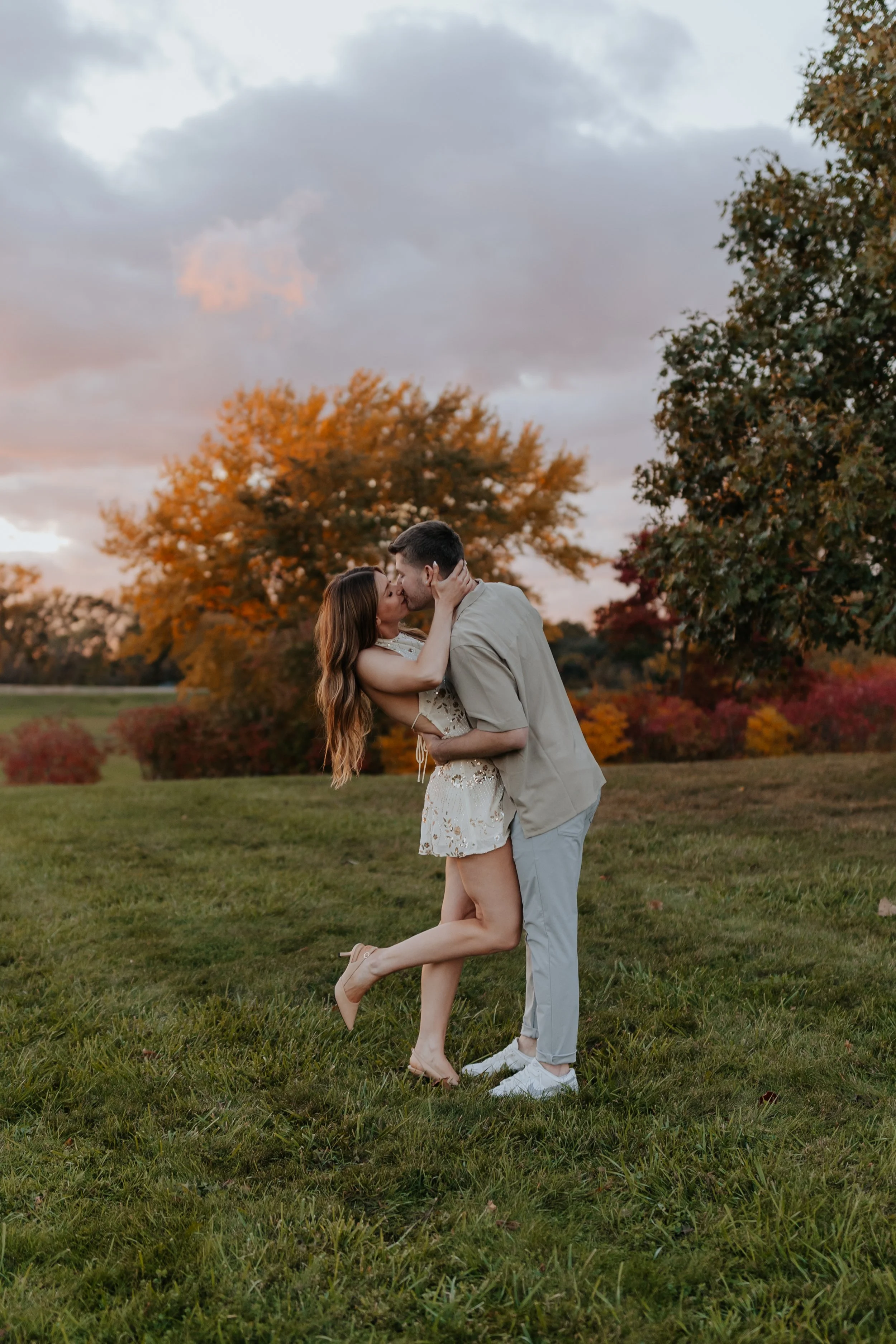 A couple kissing outdoors during autumn, with colorful trees and a cloudy sky in the background.
