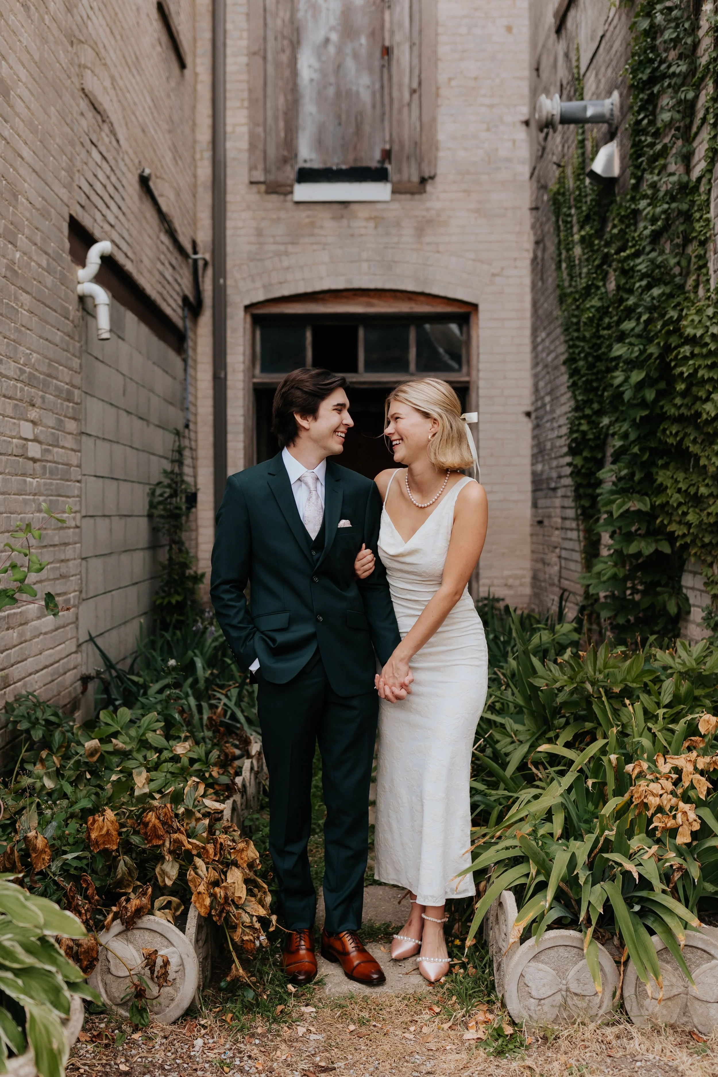A newlywed couple holding hands, smiling, and looking at each other in an outdoor setting with brick walls, greenery, and plants.