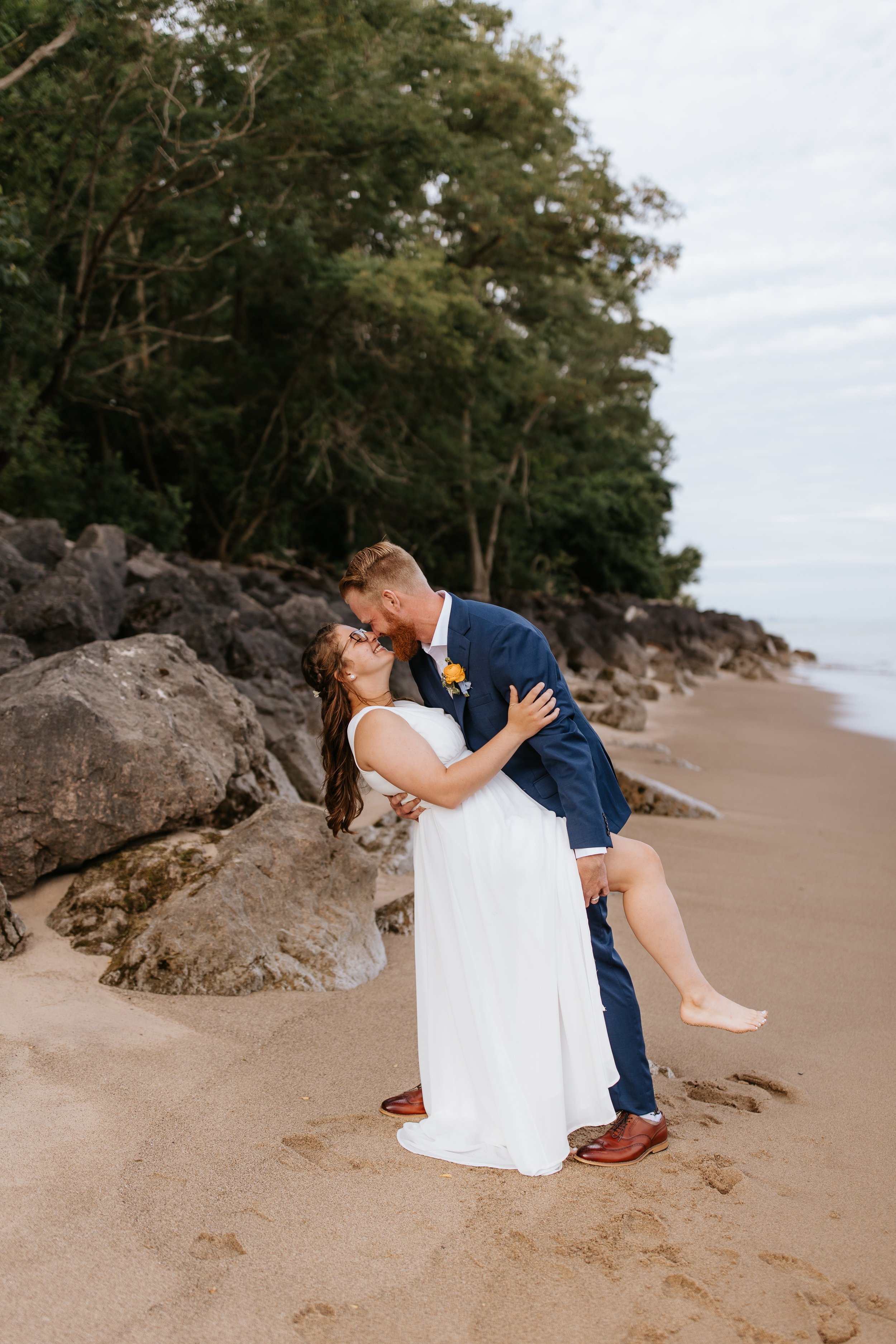 A couple dressed in wedding attire on a sandy beach, with the man dipping the woman, and trees in the background.