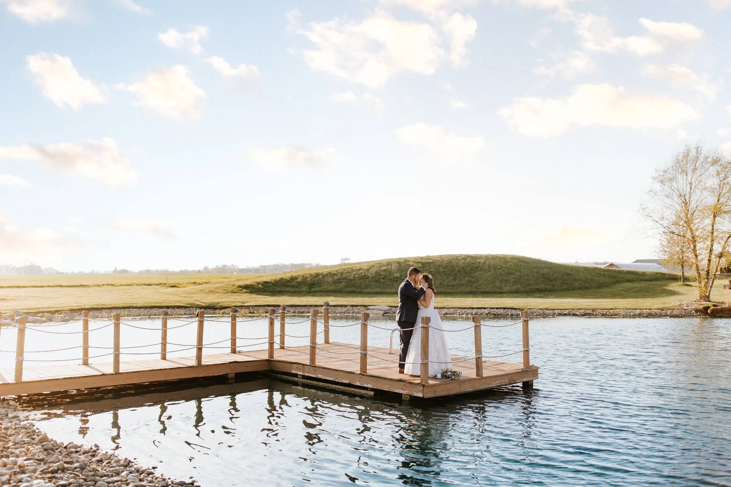 A bride and groom standing on a wooden dock by the water, sharing a kiss outdoors on a sunny day with a grassy hill, trees, and a blue sky with clouds in the background.