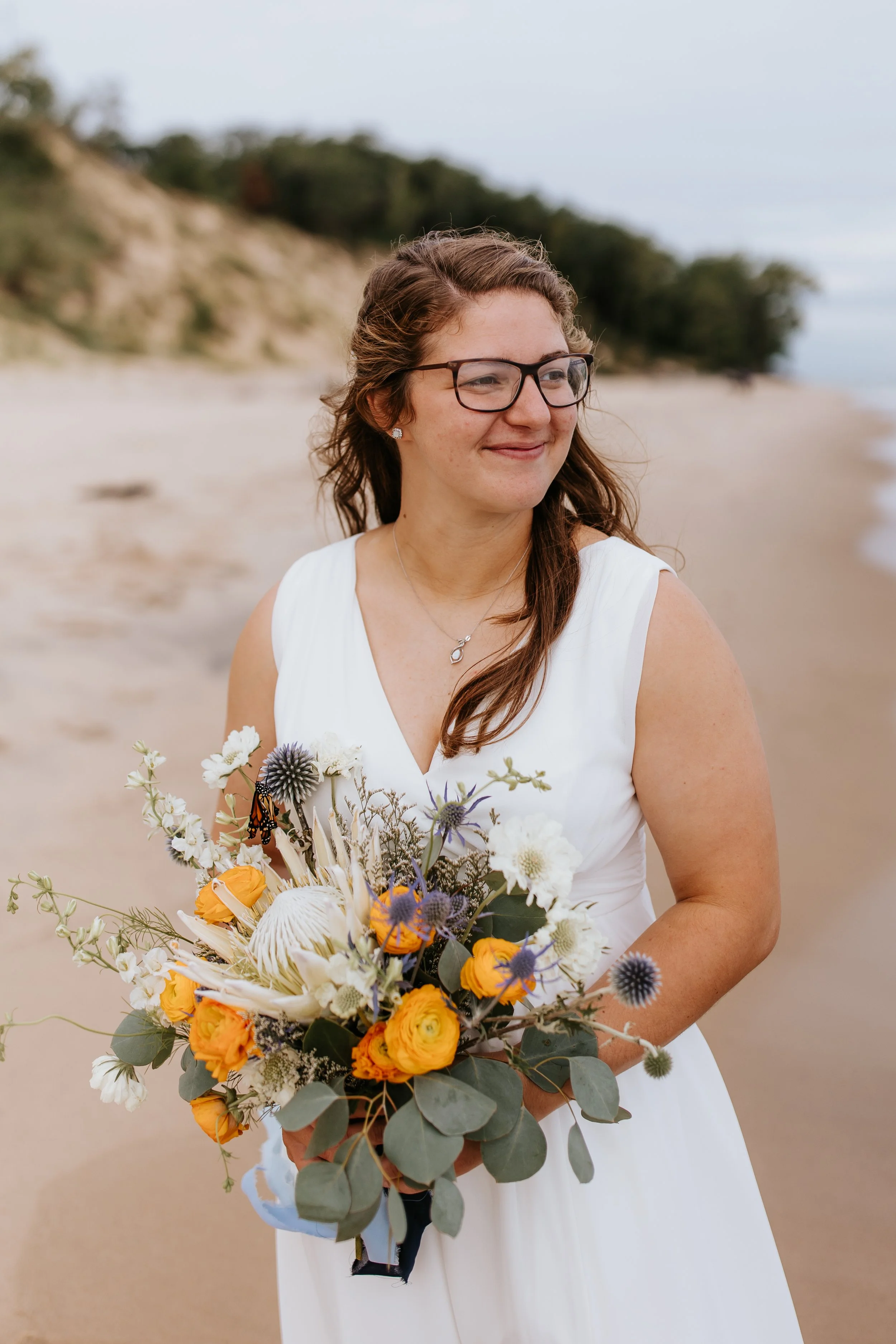 A woman in a white dress holding a bouquet of yellow, white, and purple flowers on a beach.