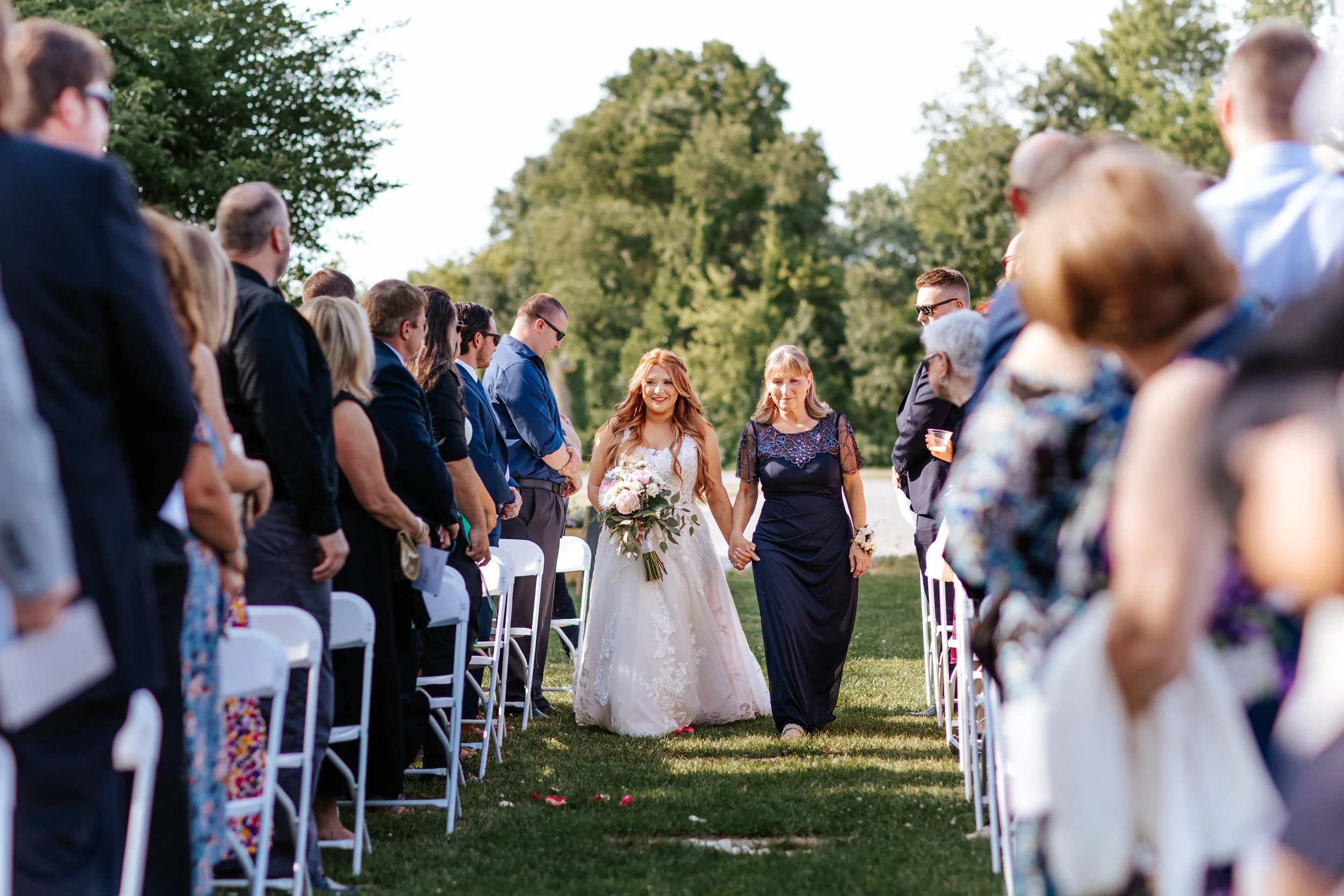 A bride holding hands with an older woman, walking down an outdoor aisle at a wedding, surrounded by seated guests.