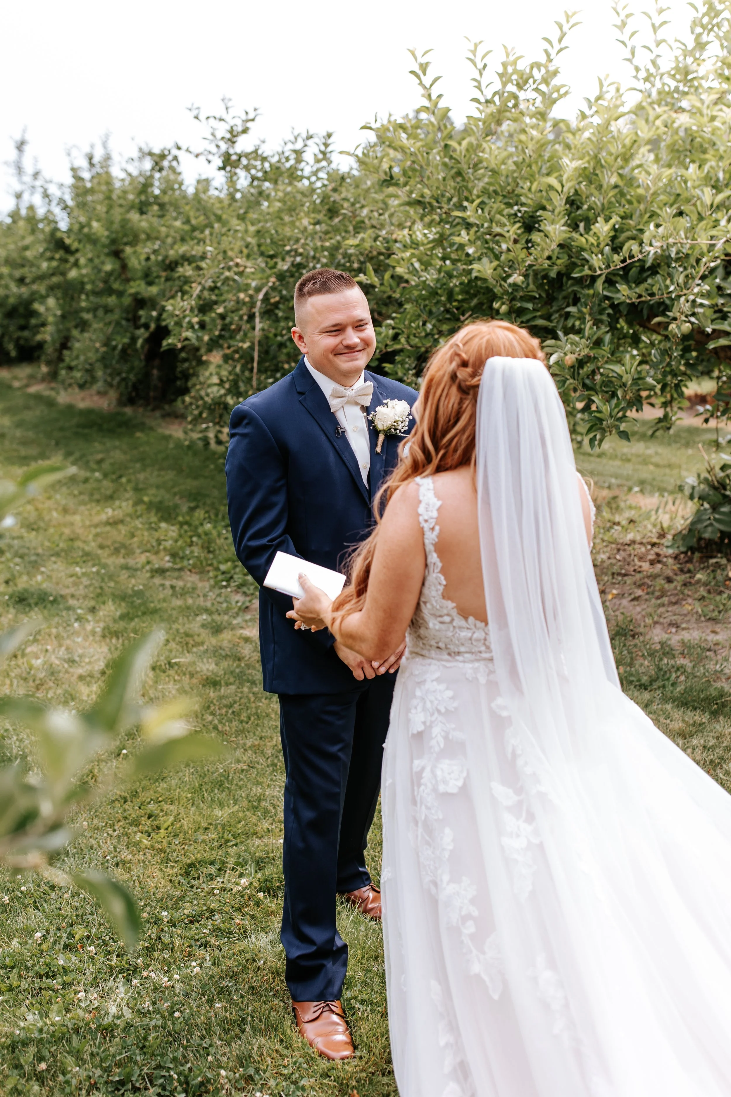 A bride and groom standing outdoors during a wedding ceremony, exchanging vows amid greenery and trees.