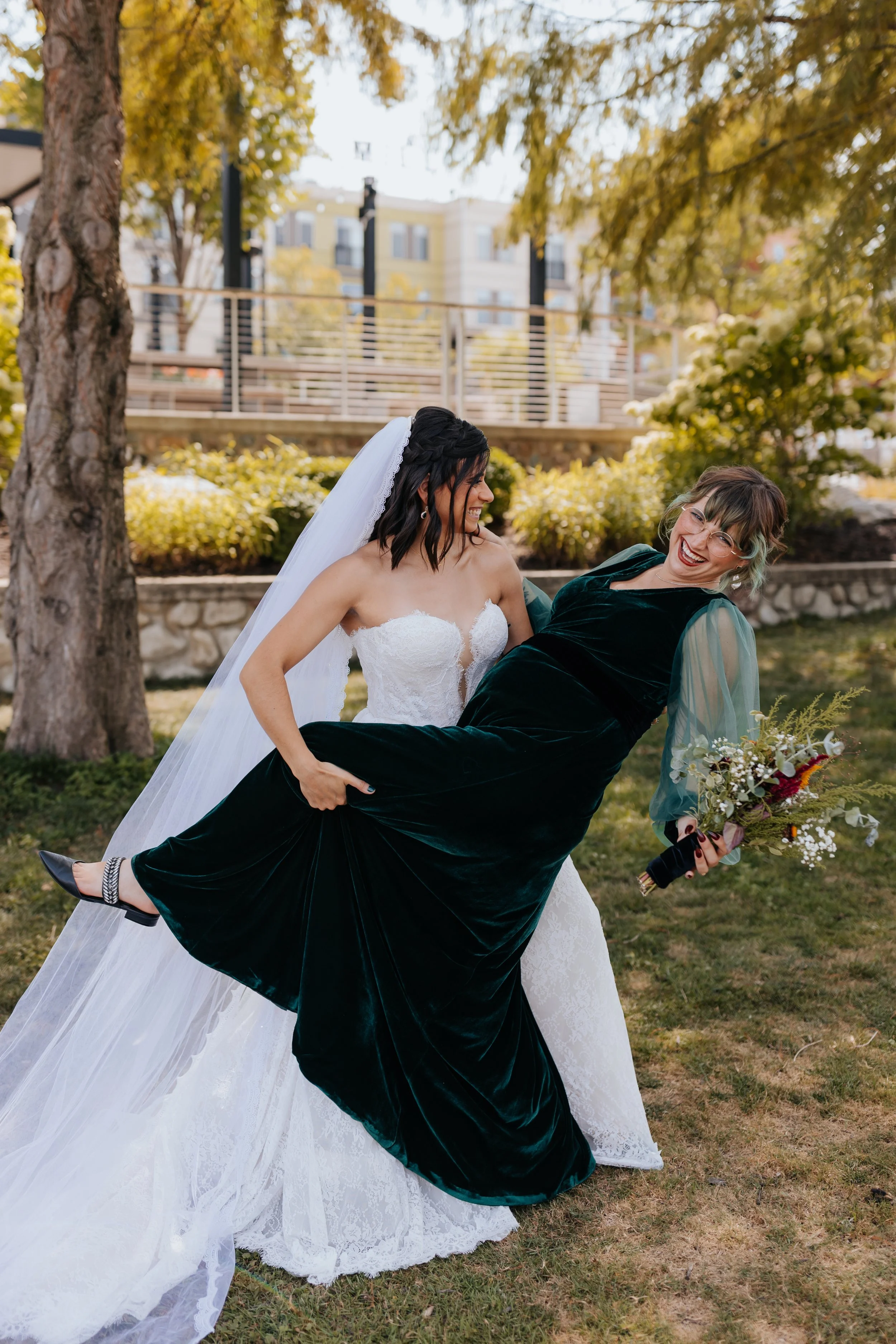 A bride and a woman, likely her partner, having fun outdoors during daytime. The bride wears a white lace wedding dress and veil, and the other woman wears a dark green velvet dress and glasses. The woman in the dark green dress is holding a bouquet 
