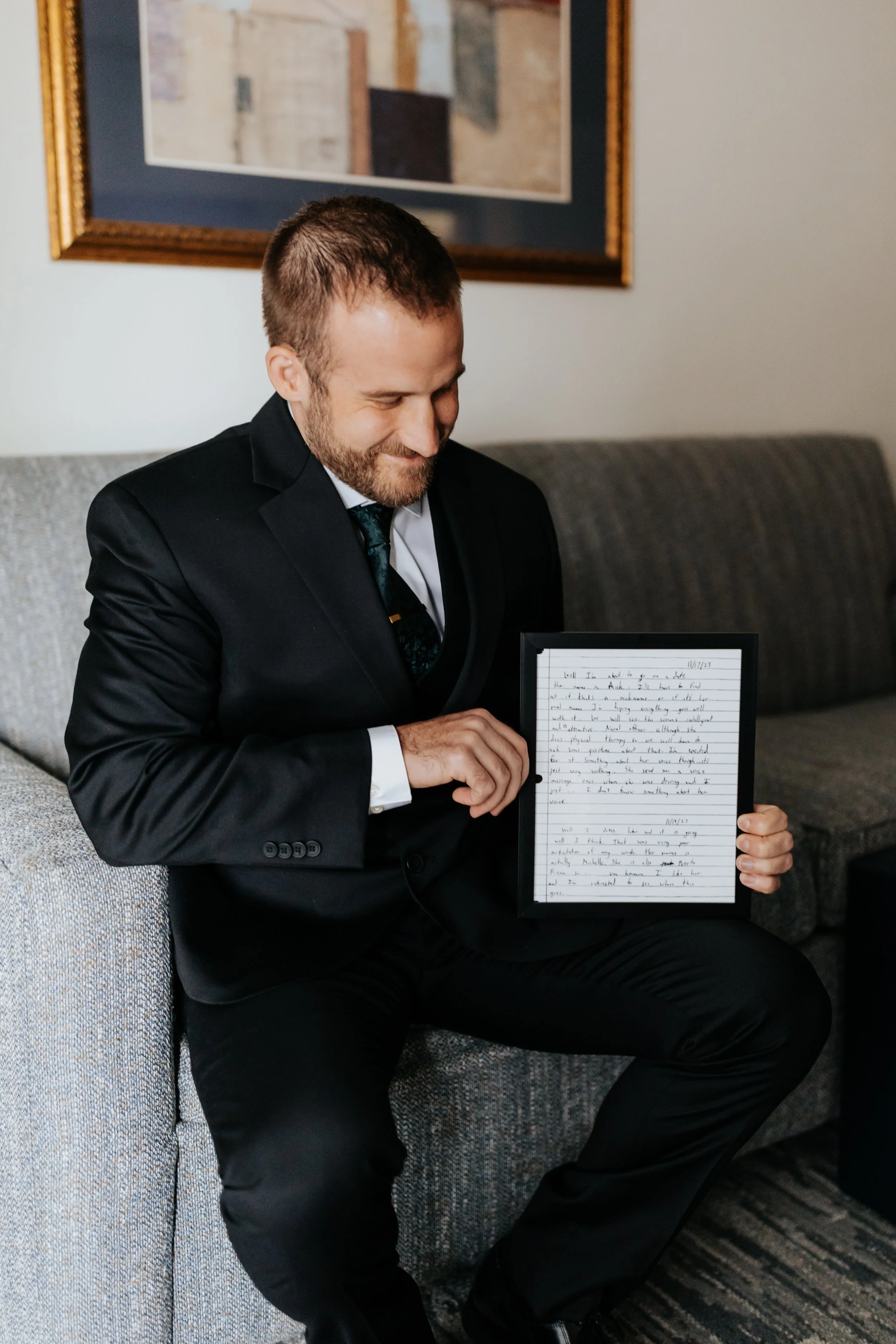 A man in a black suit sits on a gray couch, smiling while holding a framed handwritten letter.