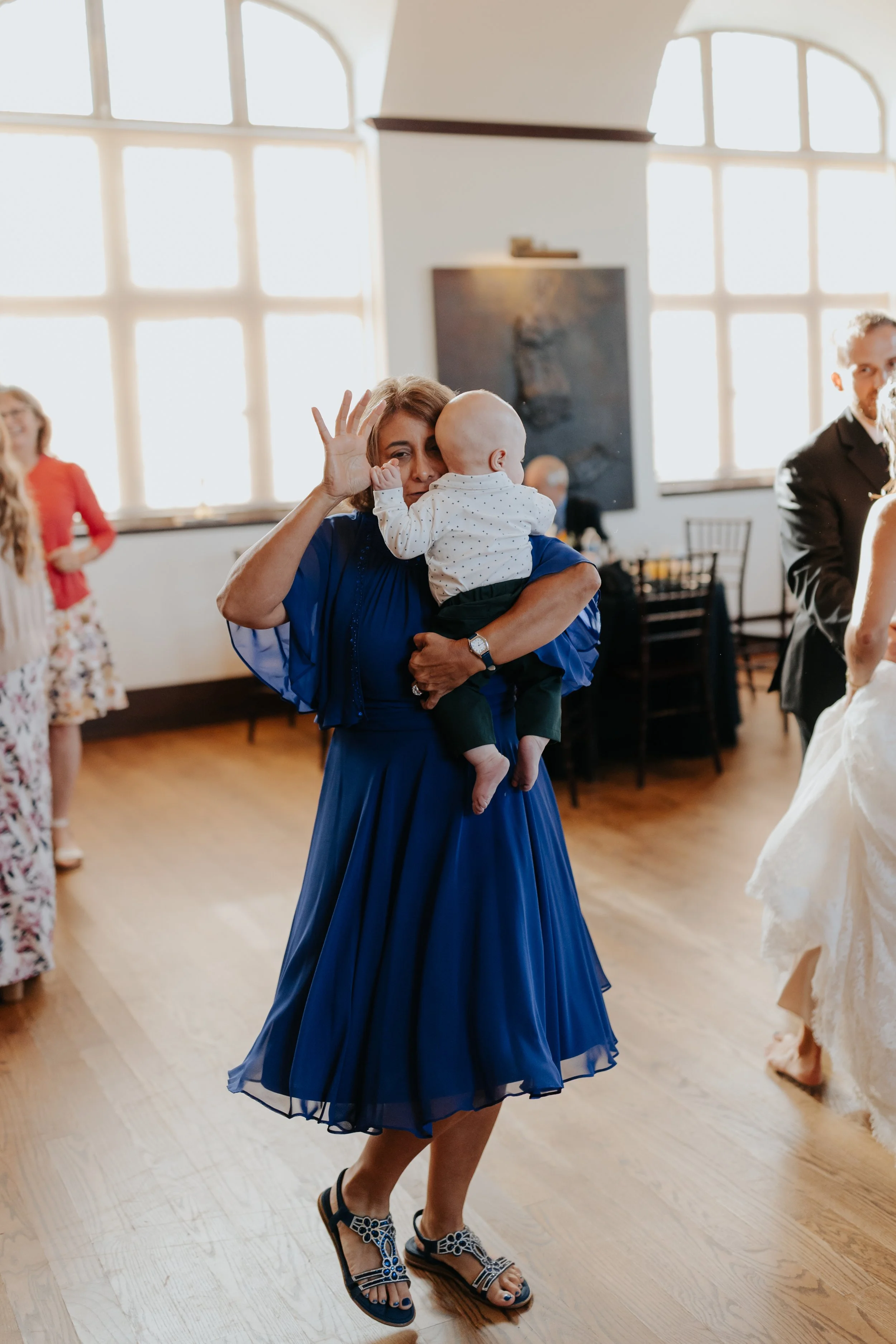 A woman wearing a blue dress and sandals holding a baby who is covering her face, at a social gathering in a room with large windows and wooden floors.