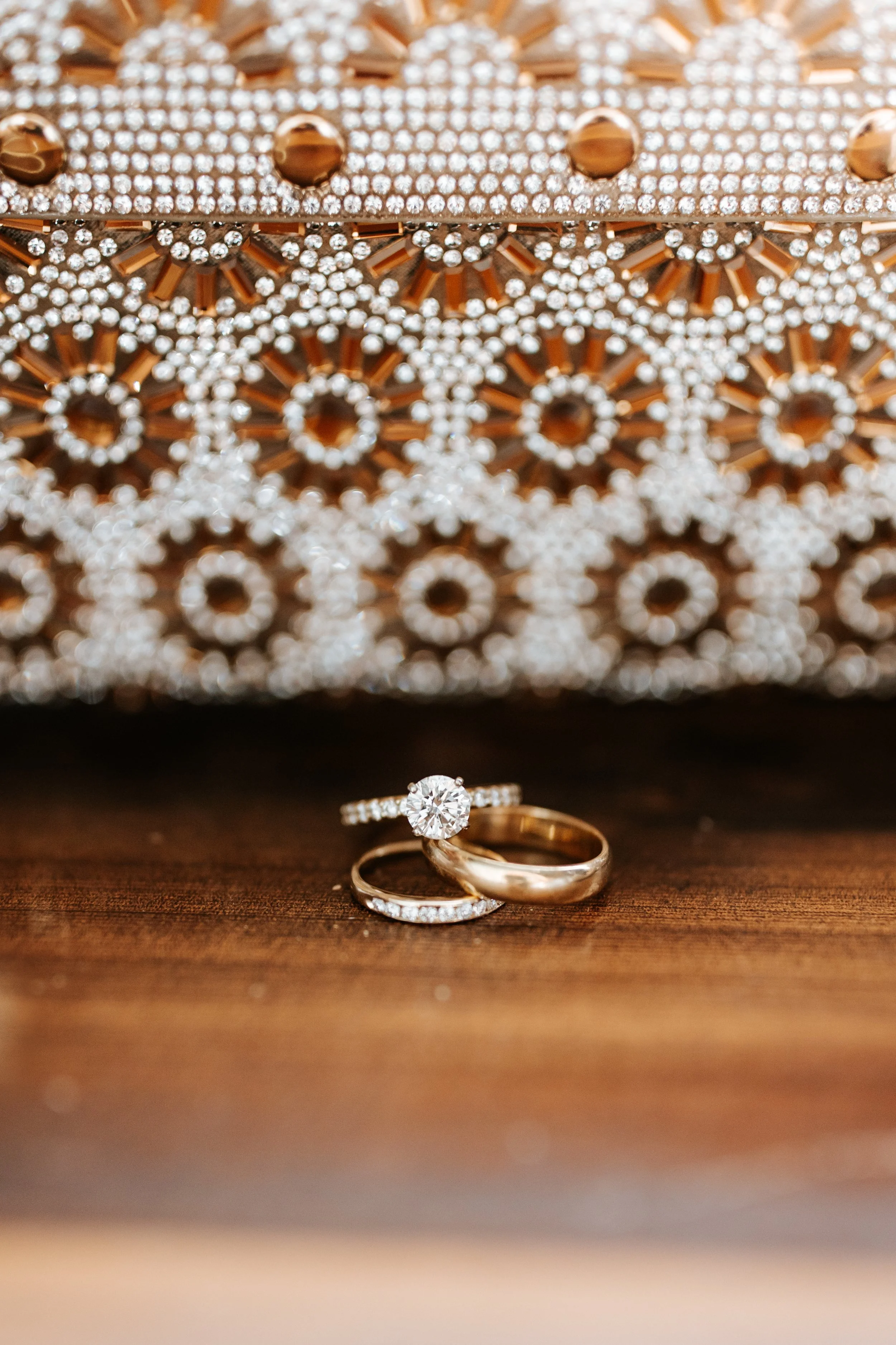 Close-up of a diamond engagement ring and wedding band on a wooden surface, with an ornate, rhinestone-encrusted clutch purse in the background.